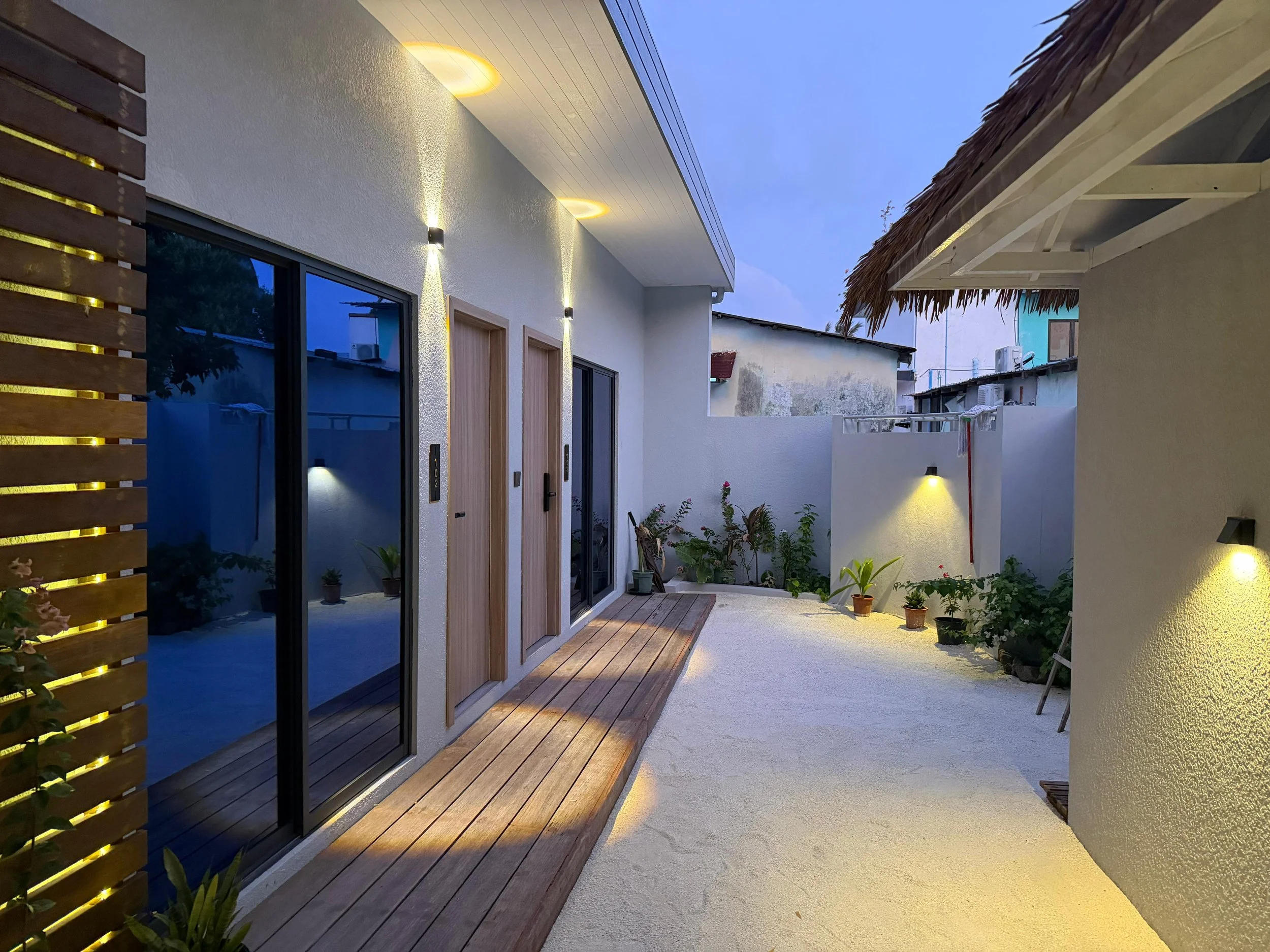 A modern outdoor patio area at dusk with sliding glass doors, potted plants, and wall-mounted lights illuminating the space.