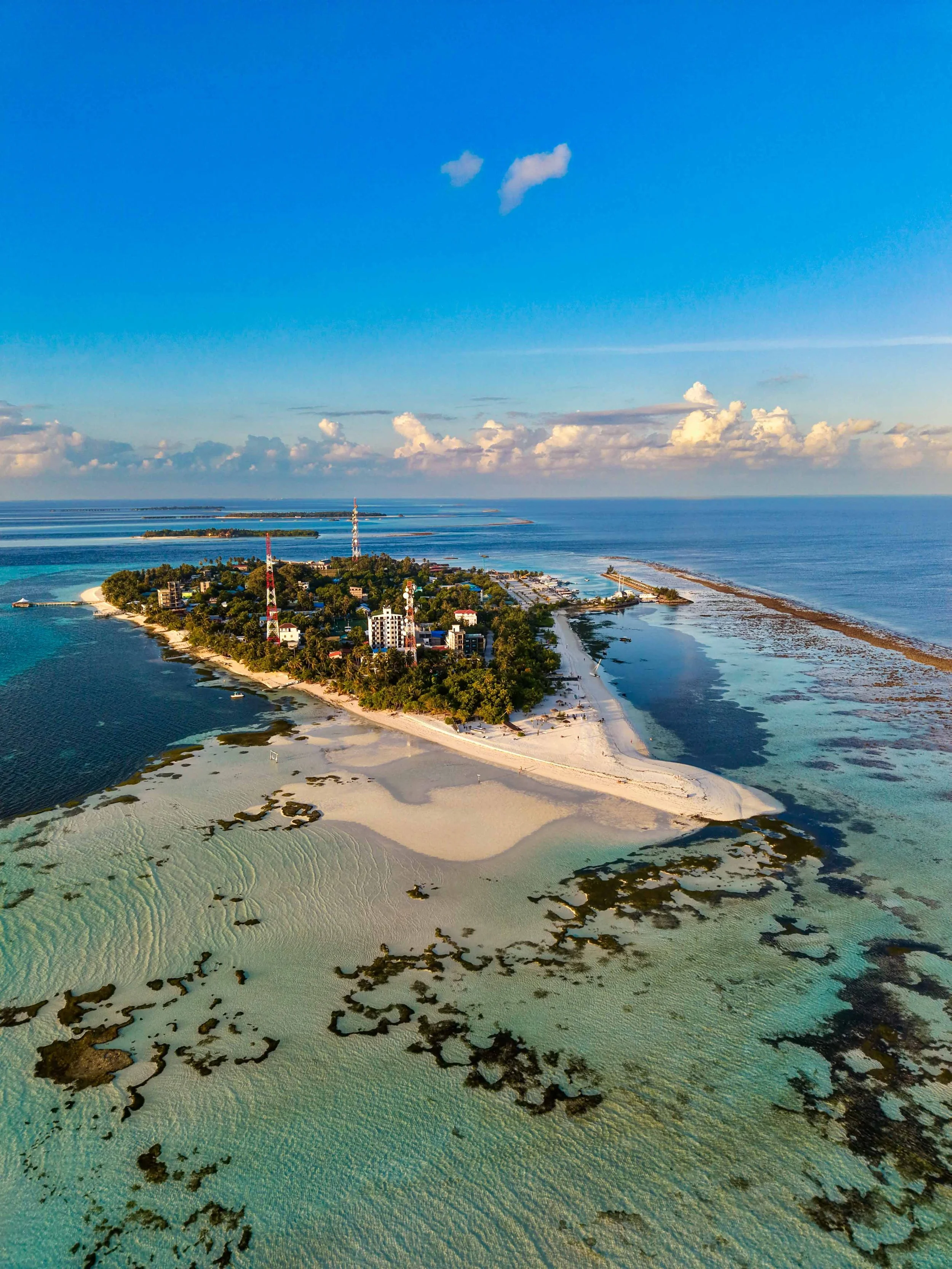 Aerial view of a tropical island with sandy beaches, turquoise waters, and lush greenery under a partly cloudy sky.