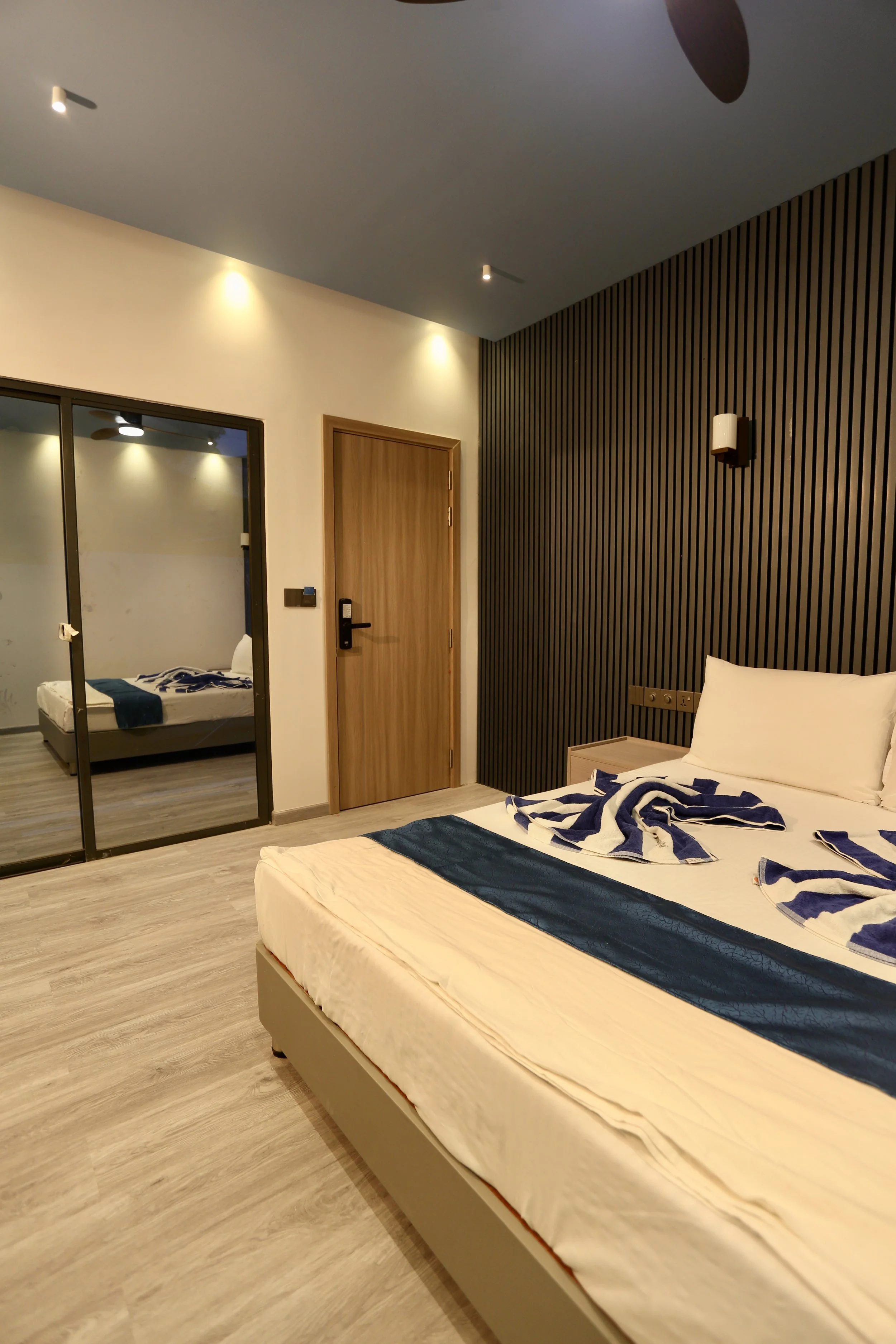 A modern hotel room with a bed covered in white and navy bedding, bedside table, and a mirrored closet. The room has wood and black vertical striped accent wall, with ceiling lights illuminating the space.