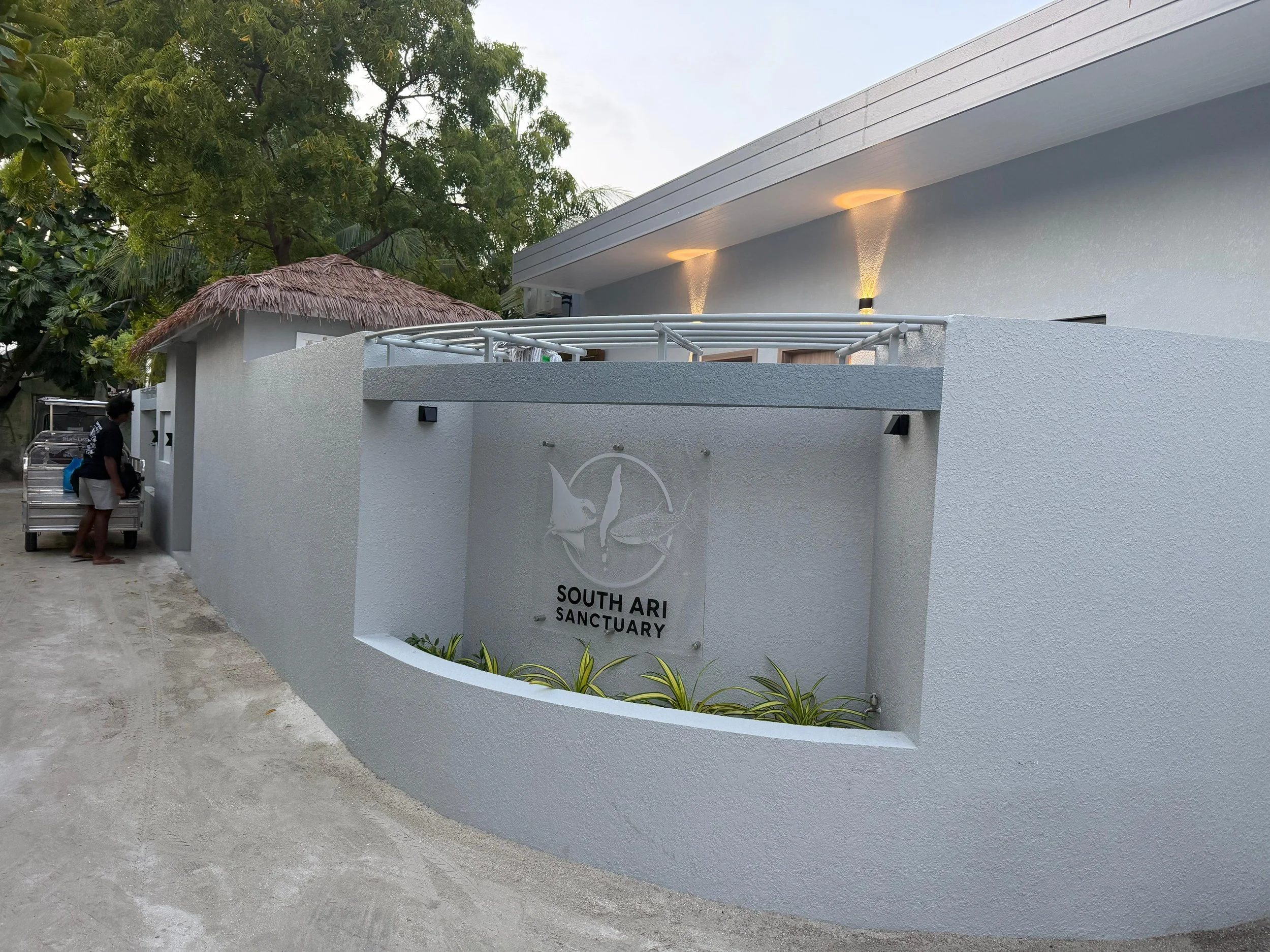 Front view of a white building with a sign that reads 'South Ari Sanctuary' featuring a pelican logo, surrounded by a curved wall with plants, a thatched roof hut, and trees in the background.