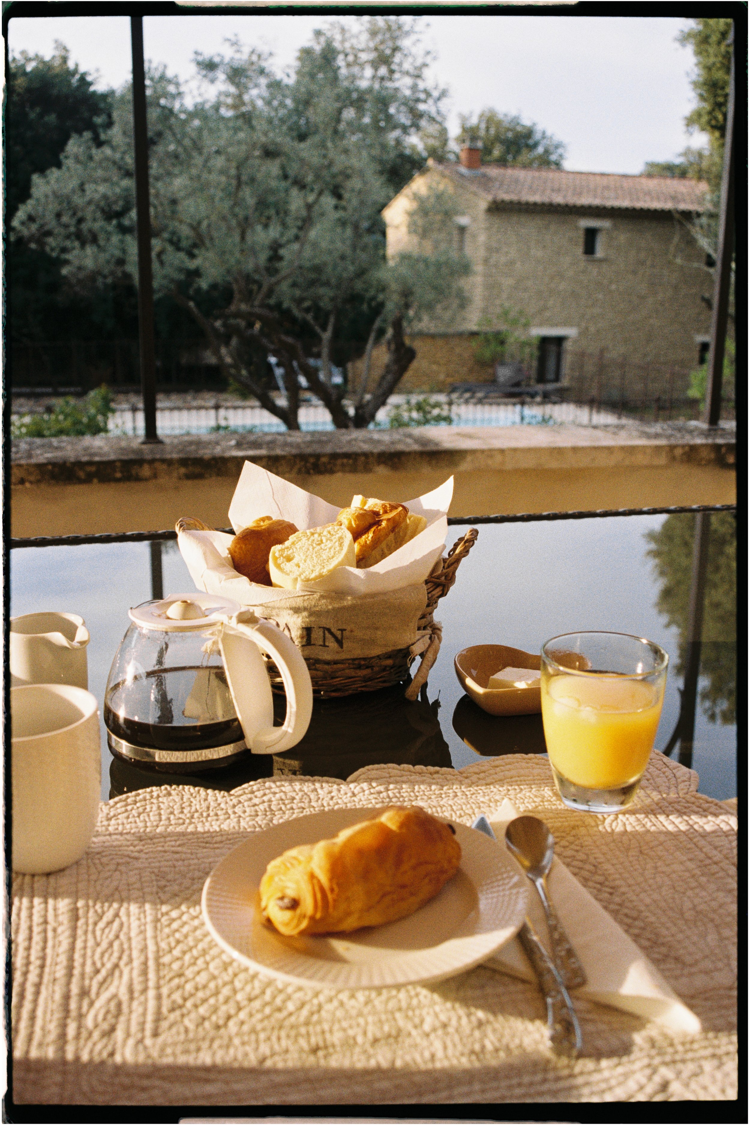 Breakfast on a table with a croissant, coffee, orange juice, a basket of bread, and creamer, with a pond and house in the background. Photographed by Emily Gould, Edinburgh based photographer working across both film and digital formats.