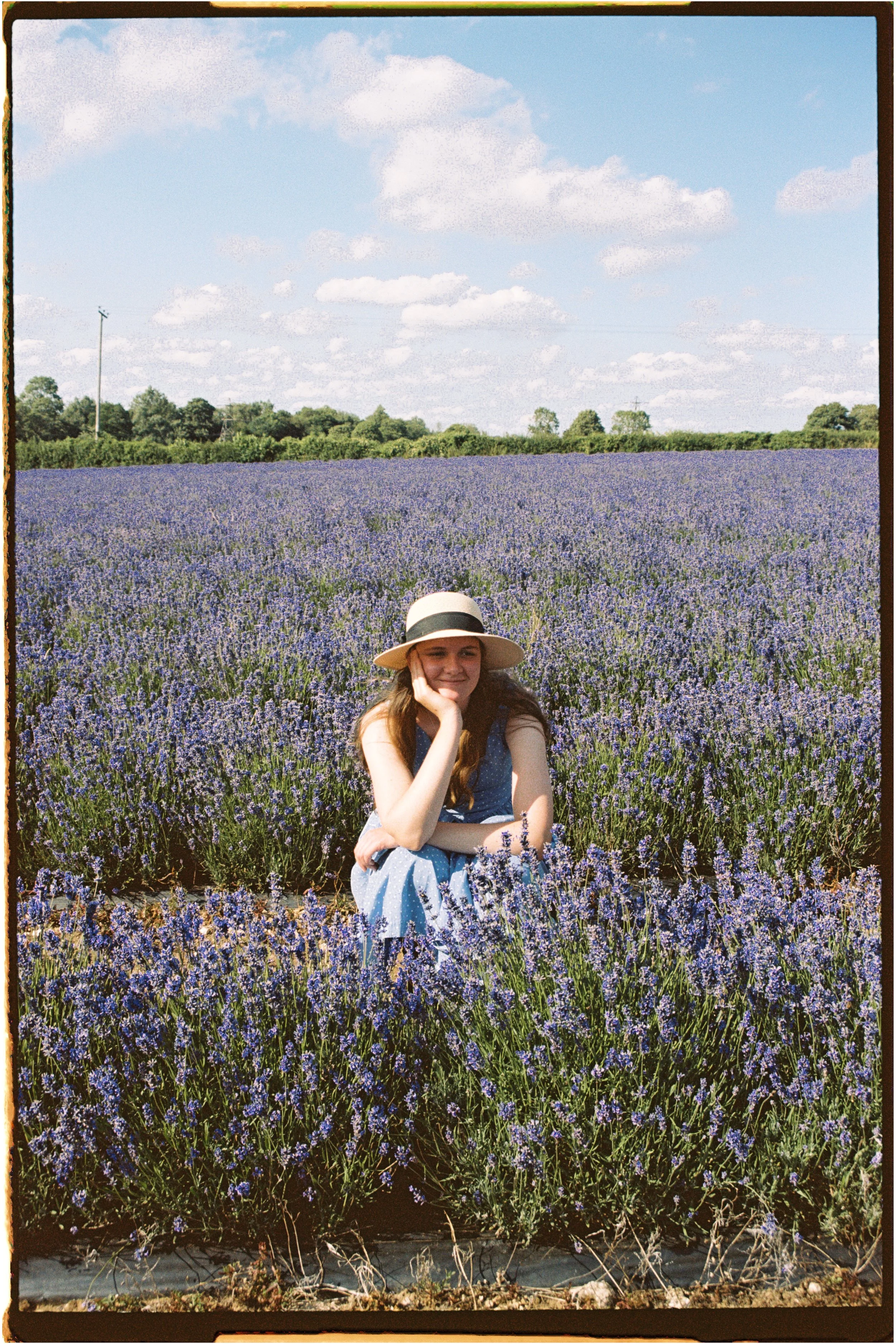 A young woman sitting in a lavender field under a blue sky with white clouds, wearing a straw hat and a blue dress. Photographed by Emily Gould, Edinburgh based photographer working across both film and digital formats.