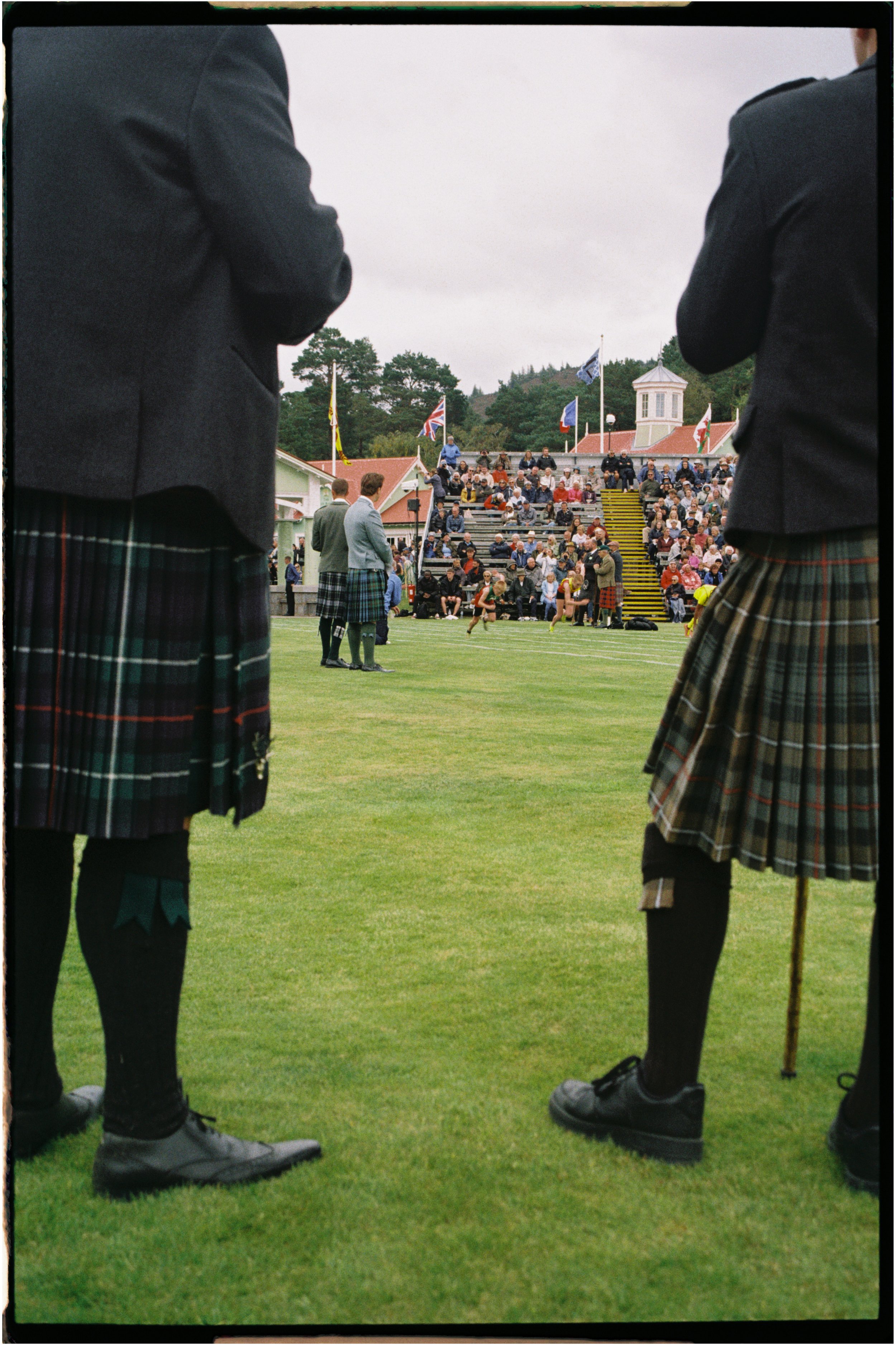 People wearing traditional Scottish kilts watching a sporting event on a grassy field with spectators in the stands and flags in the background. Photographed by Emily Gould, Edinburgh based photographer working across both film and digital formats.