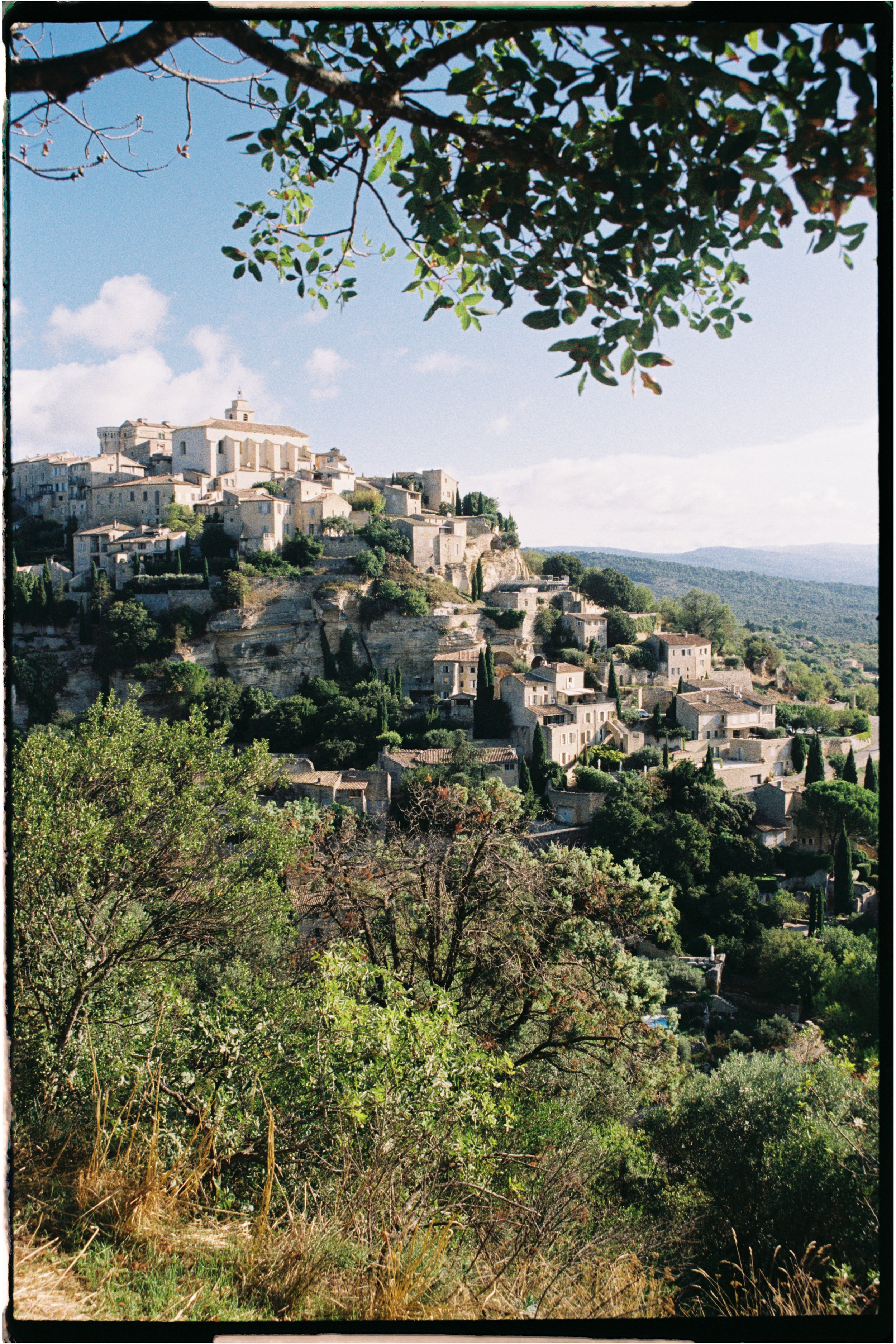 A hillside Provence village with white buildings, tall trees, and a partly cloudy sky, photographed by Emily Gould, Edinburgh based photographer working across both film and digital formats.