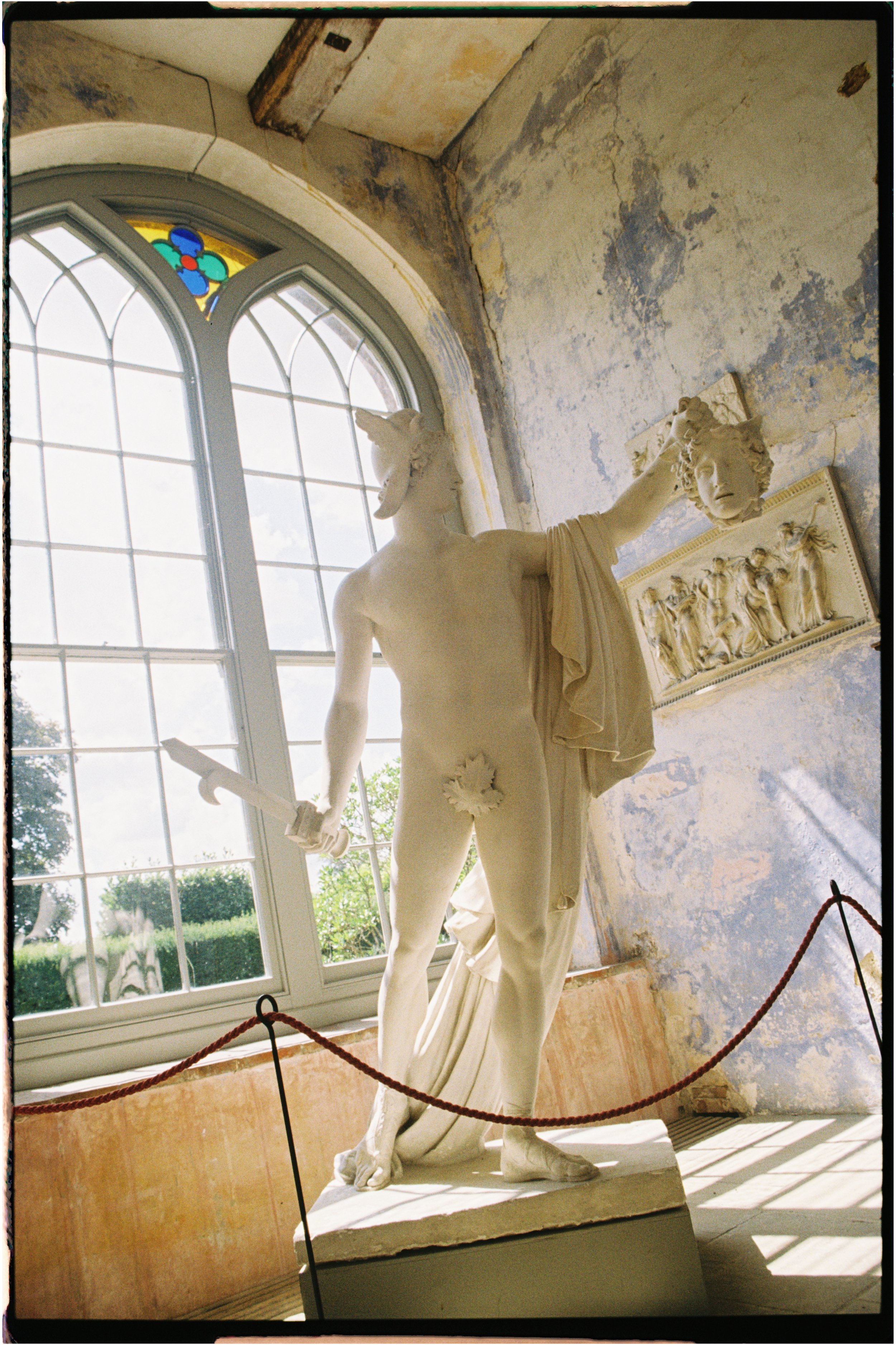 A marble sculpture of a mythical creature with a human body and a unicorn head, holding a mask in one hand and a sword in the other, displayed indoors near a large window with stained glass at the top. Photographed by Emily Gould, Edinburgh based pho