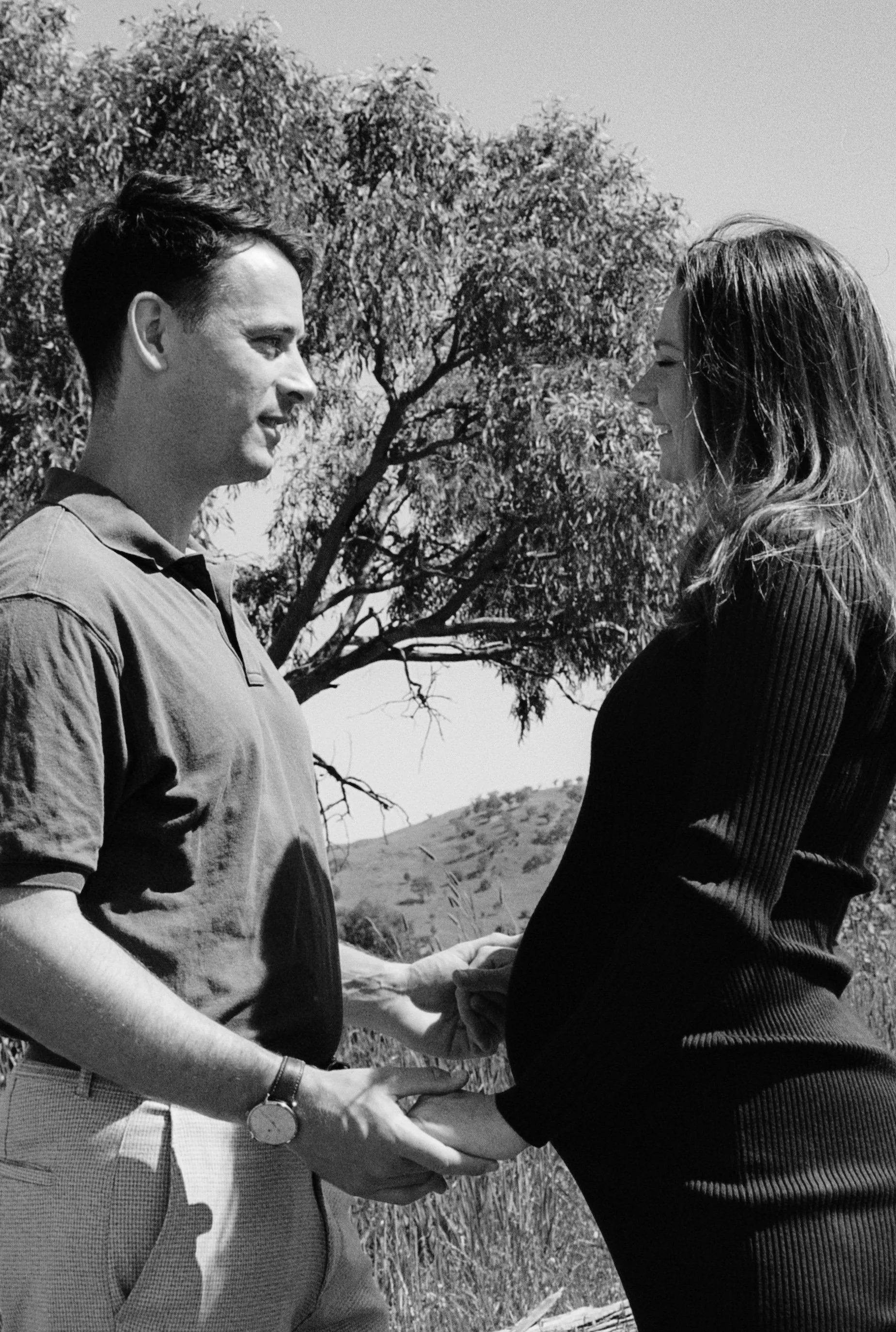 Pregnancy shoot - A man and woman holding hands outdoors, facing each other and smiling, with trees and hills in the background, in black and white. Photographed by Emily Gould, Edinburgh based photographer working across both film and digital format