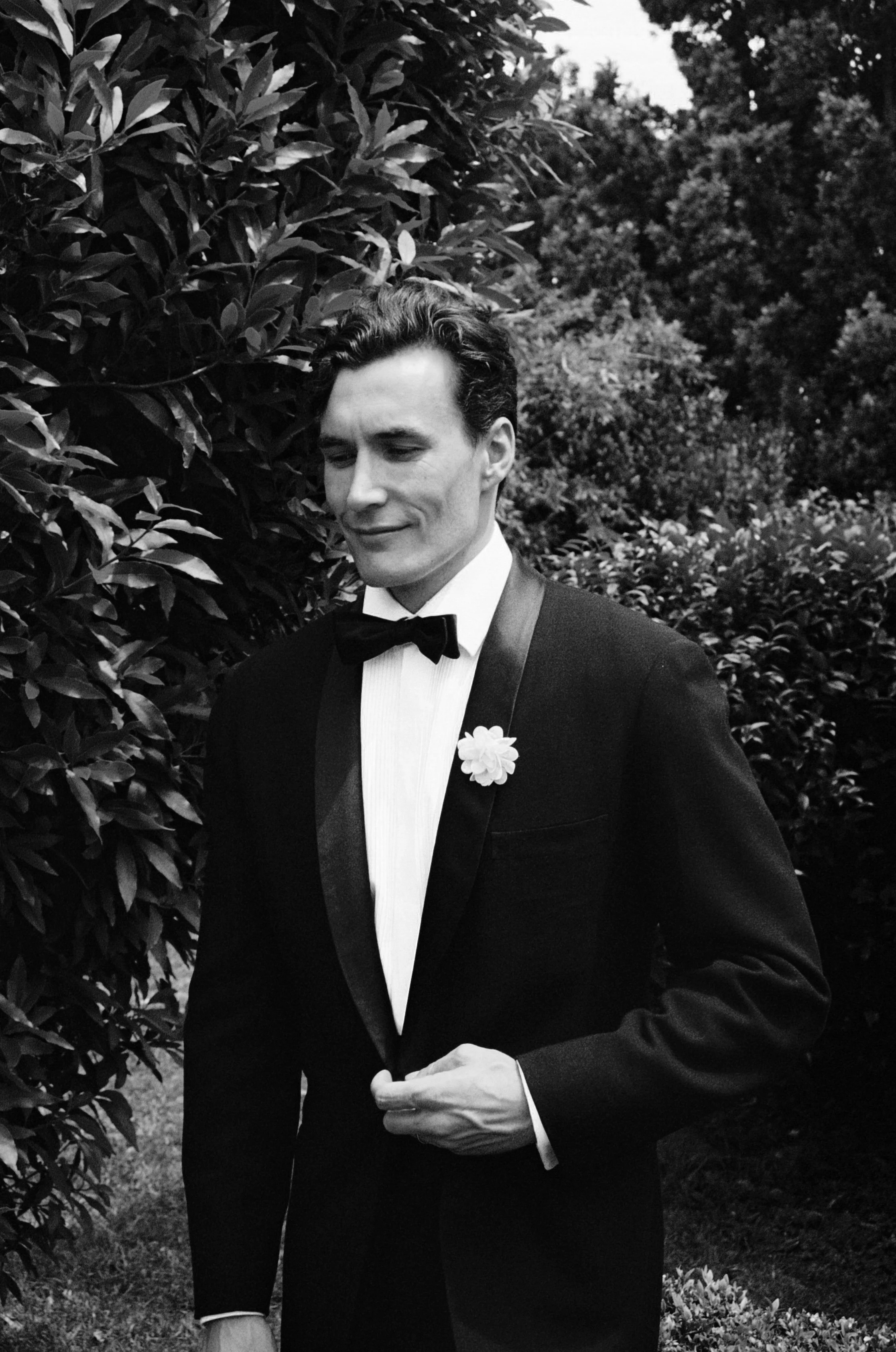 A man in formal attire, wearing a tuxedo with a bow tie, standing outdoors with trees and bushes in the background. Photographed by Emily Gould, Edinburgh based photographer working across both film and digital formats.