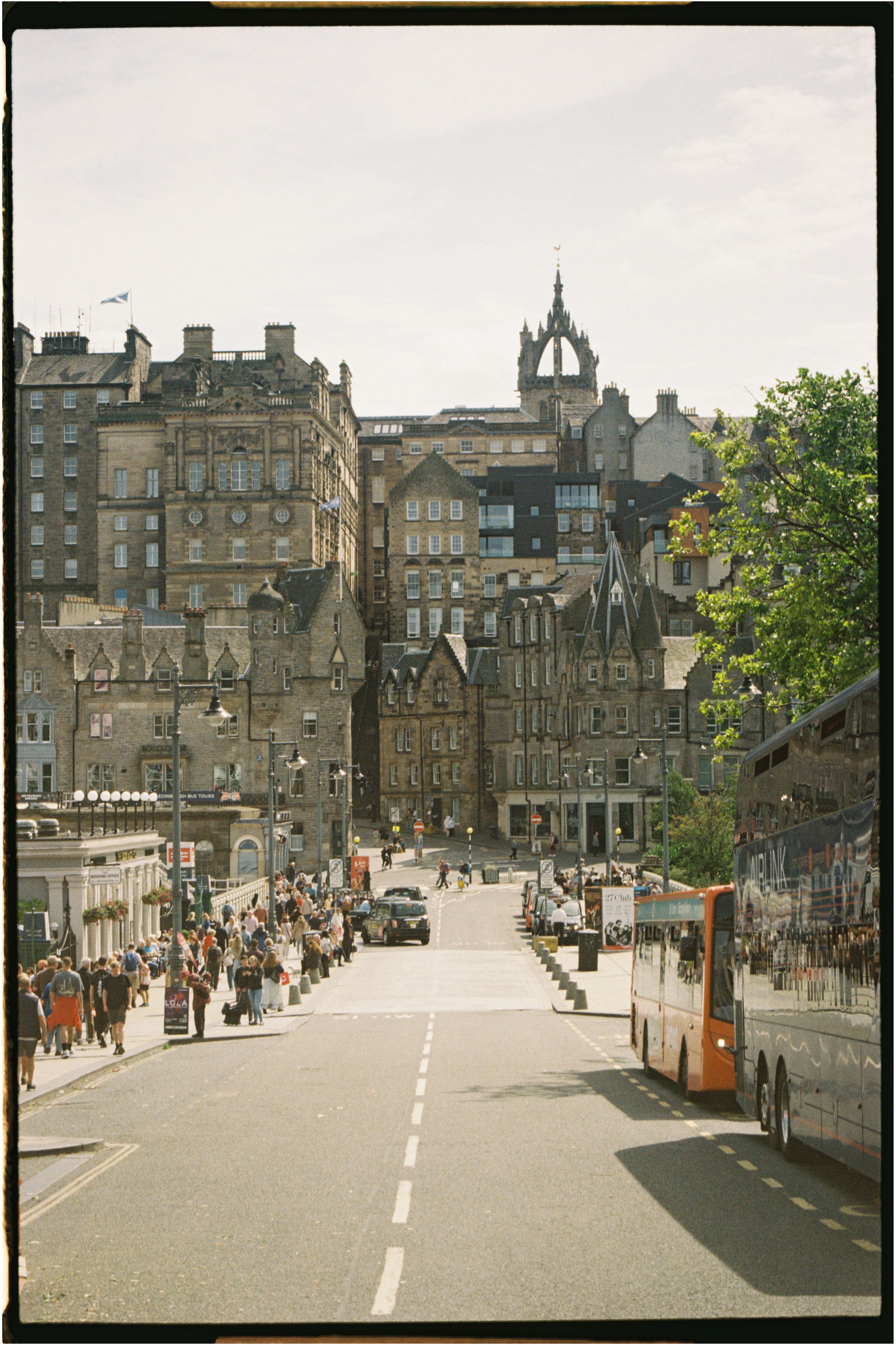 View of Edinburgh  with historic stone buildings, parked cars, a bus, pedestrians, and a prominent clock tower in the background. Photographed by Emily Gould, Edinburgh based photographer working across both film and digital formats.