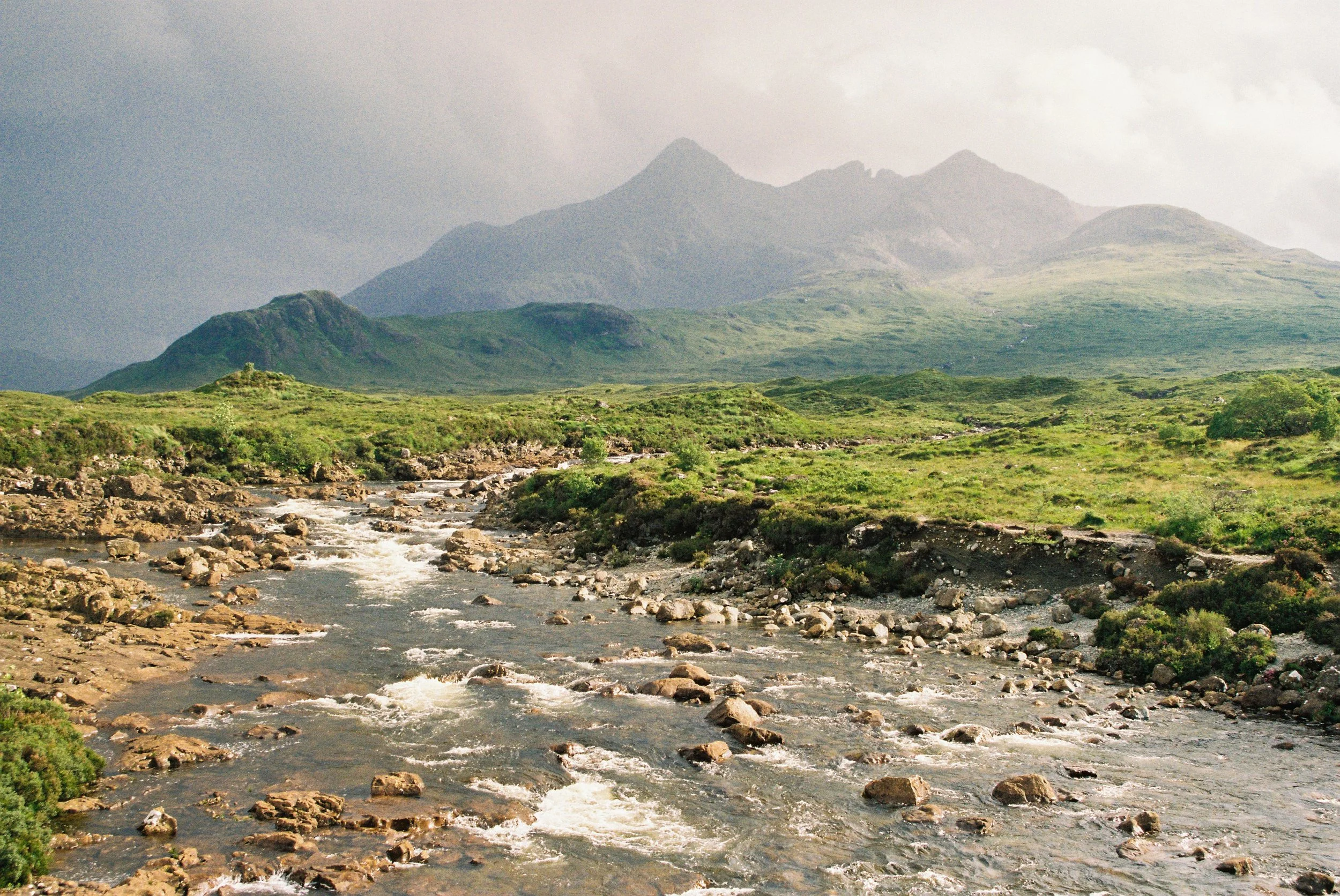 A Highlands landscape with a rocky stream in the foreground and green grassy hills leading up to distant mountains
