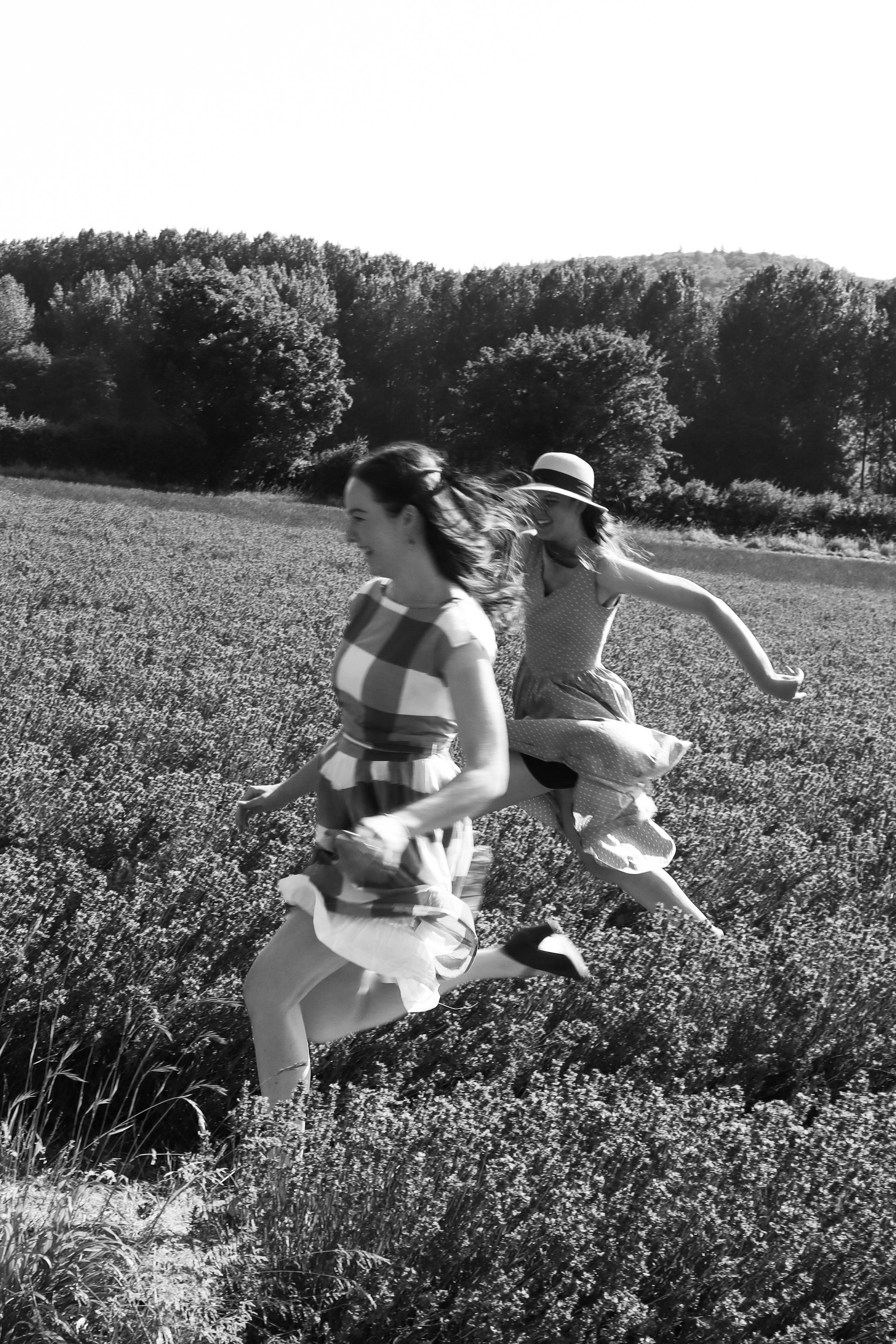 Two women running through a field of flowers, smiling and enjoying themselves, with trees and hills in the background. Photographed by Emily Gould, Edinburgh based photographer working across both film and digital formats.