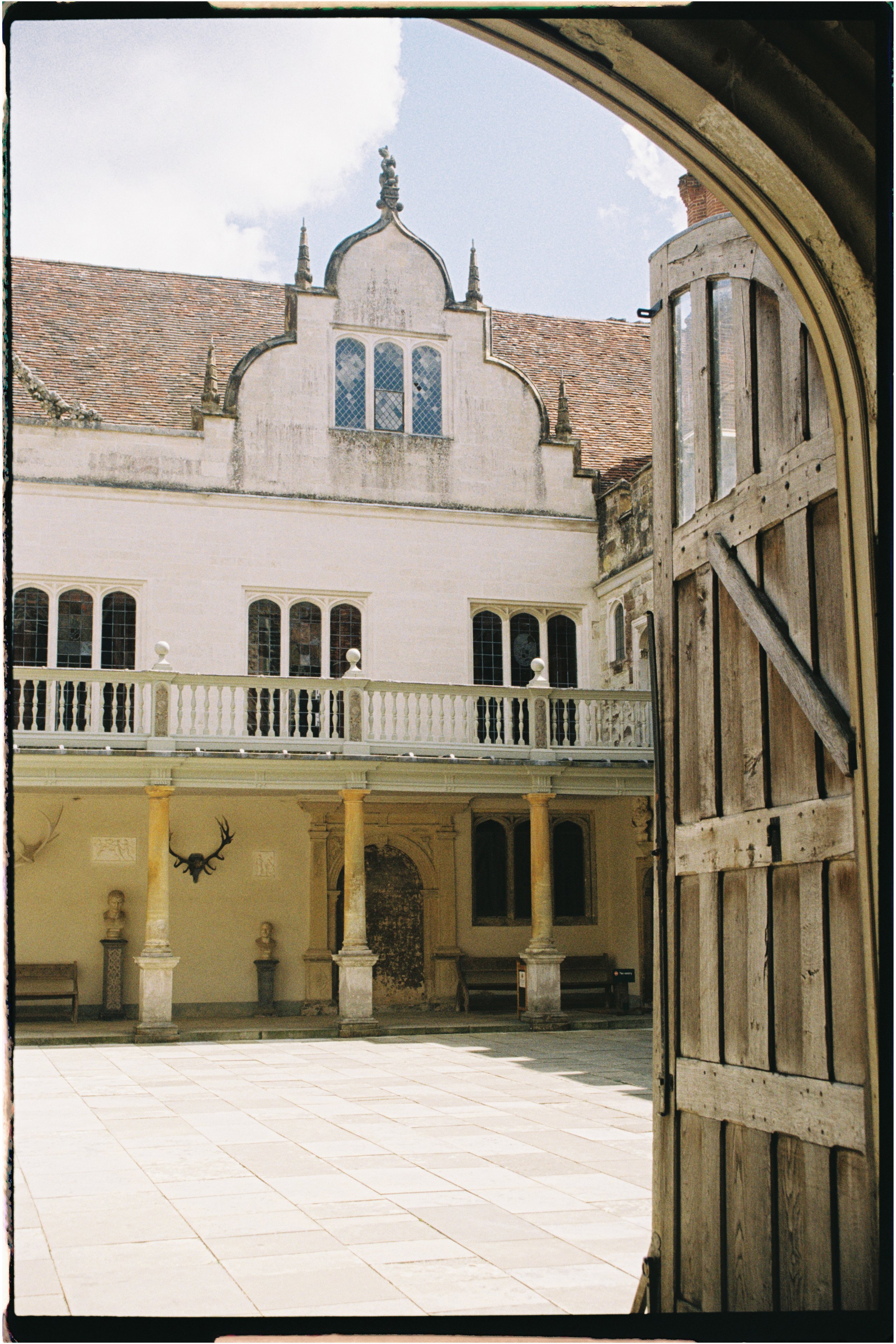 View of a historic courtyard with stone columns, benches, and a mounted deer head, seen through an open wooden gate with a historic building. Photographed by Emily Gould, Edinburgh based photographer working across both film and digital formats.