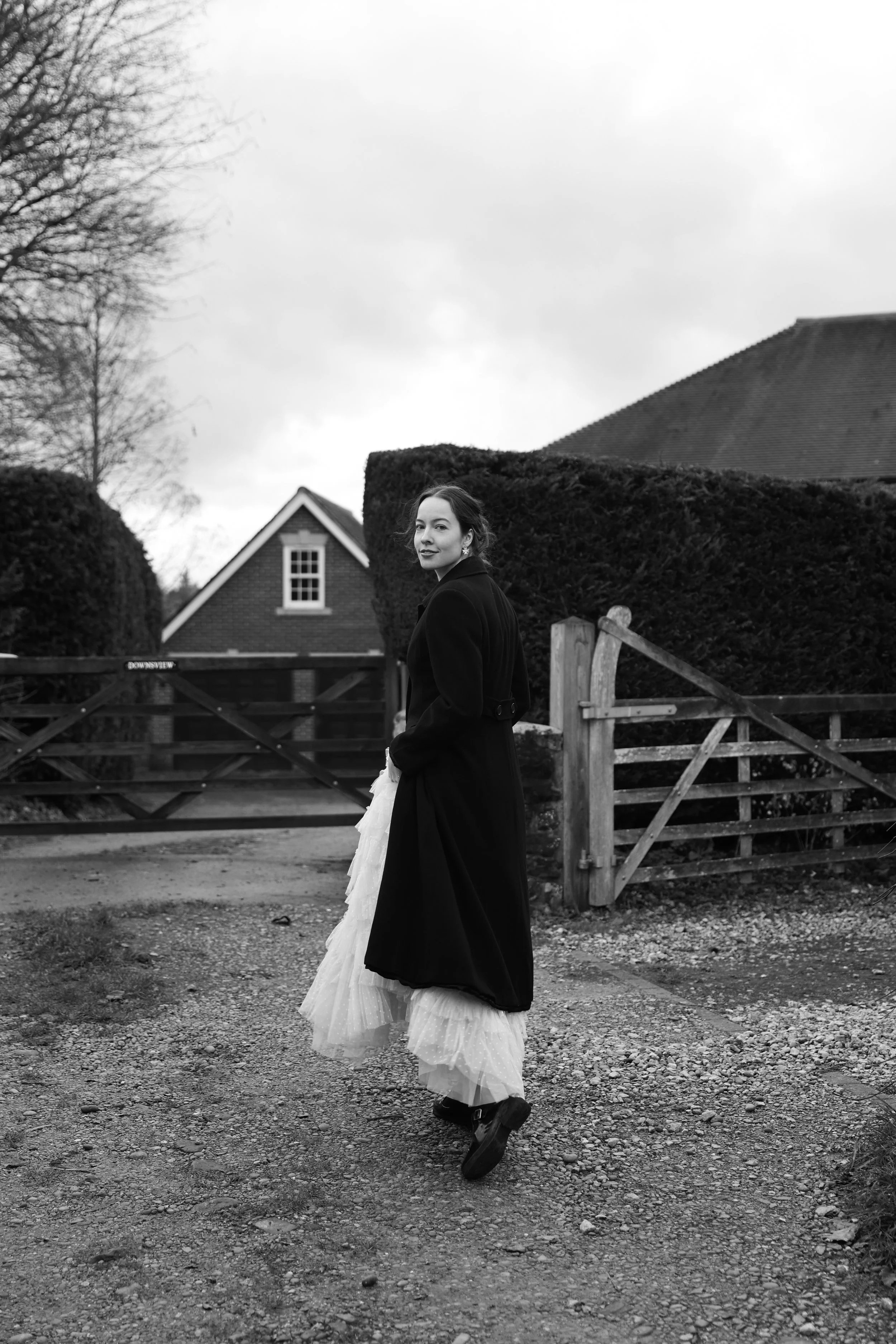 A woman in a black coat and dress standing outdoors on a gravel path, with a house, trees, and a wooden fence in the background. Photographed by Emily Gould, an Edinburgh based photographer working across film and digital formats.