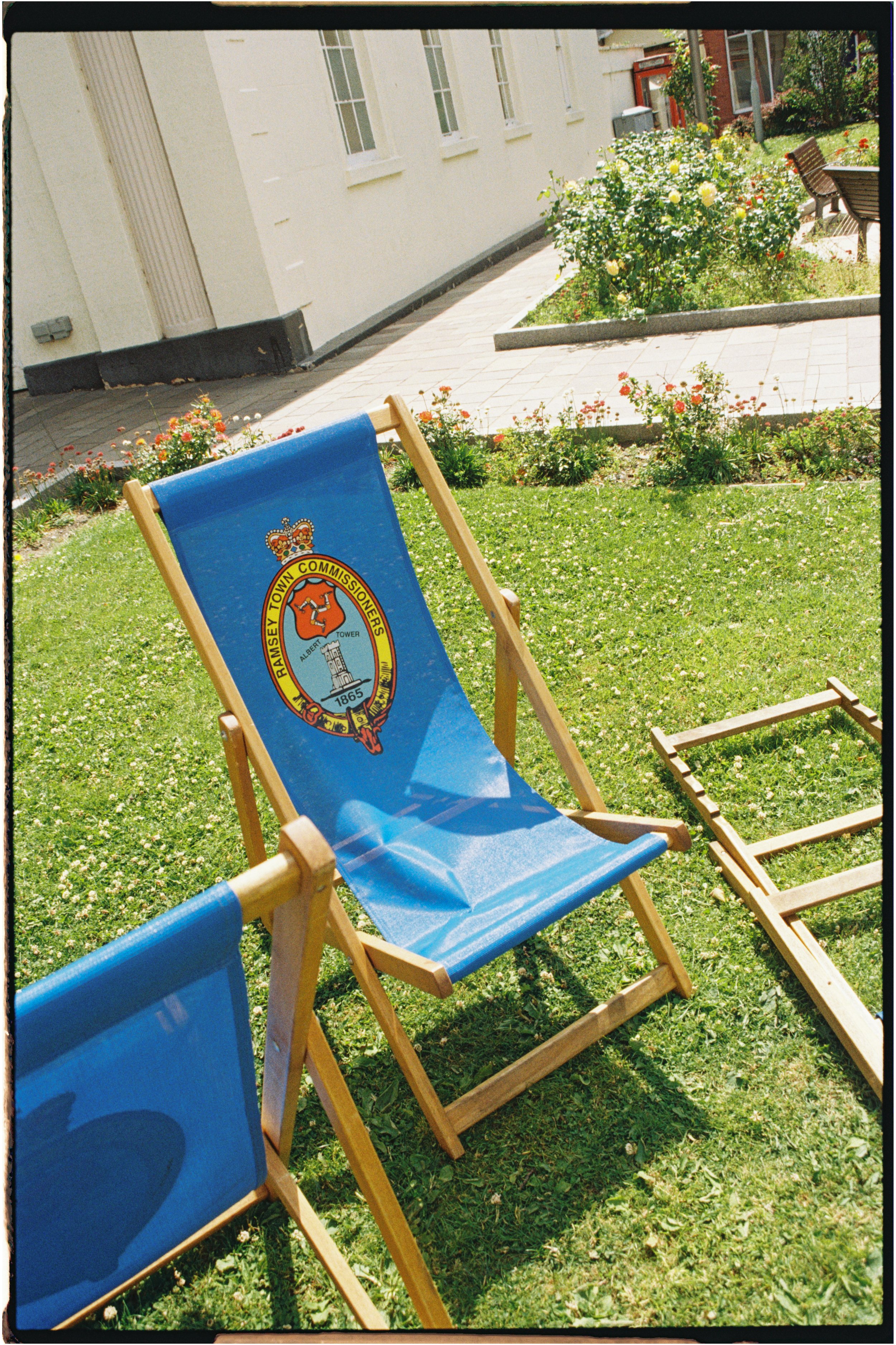 A blue collapsible chair with the Ramsey Town Commissioners logo, placed on a grassy area near flowers, with a paved walkway and building in the background. Photographed by Emily Gould, Edinburgh based photographer working across both film and digita