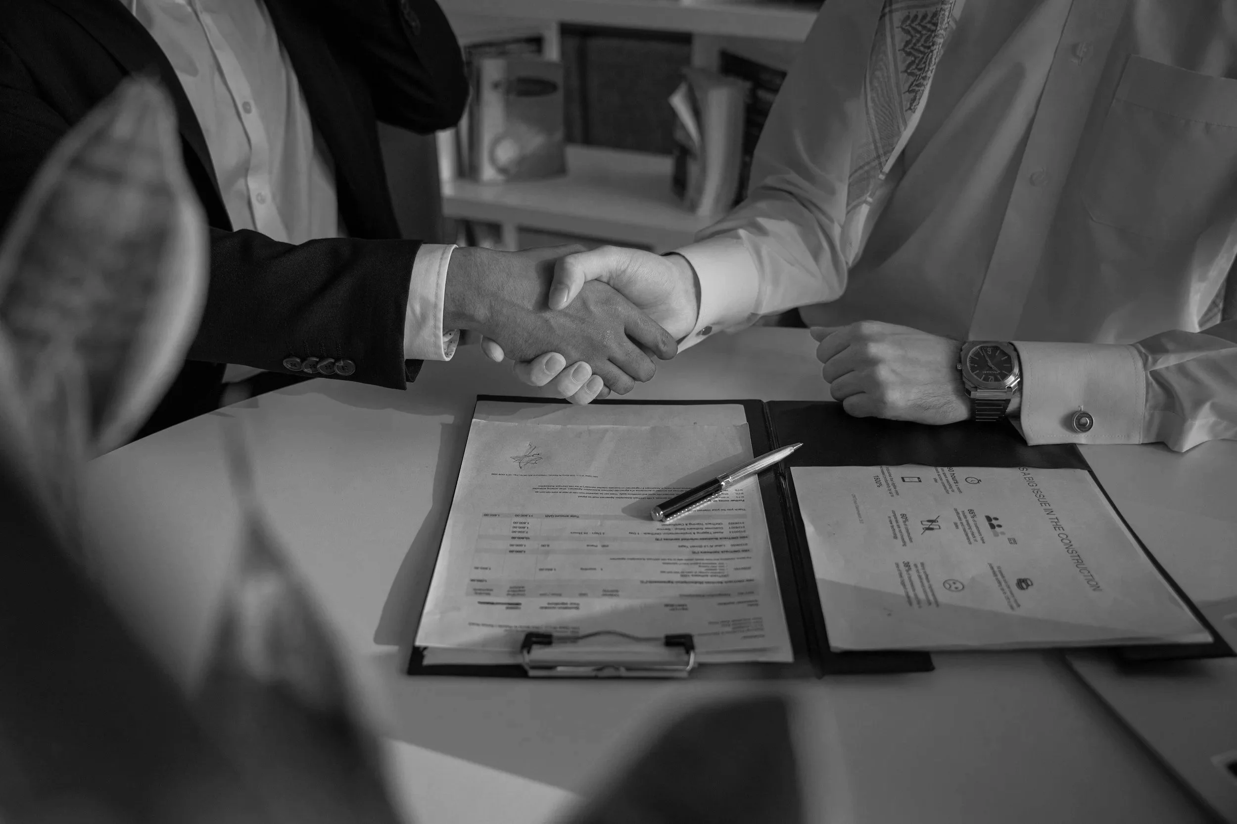 Two individuals shaking hands over a table with documents, a pen, and a certificate, suggesting a formal agreement or contract signing.