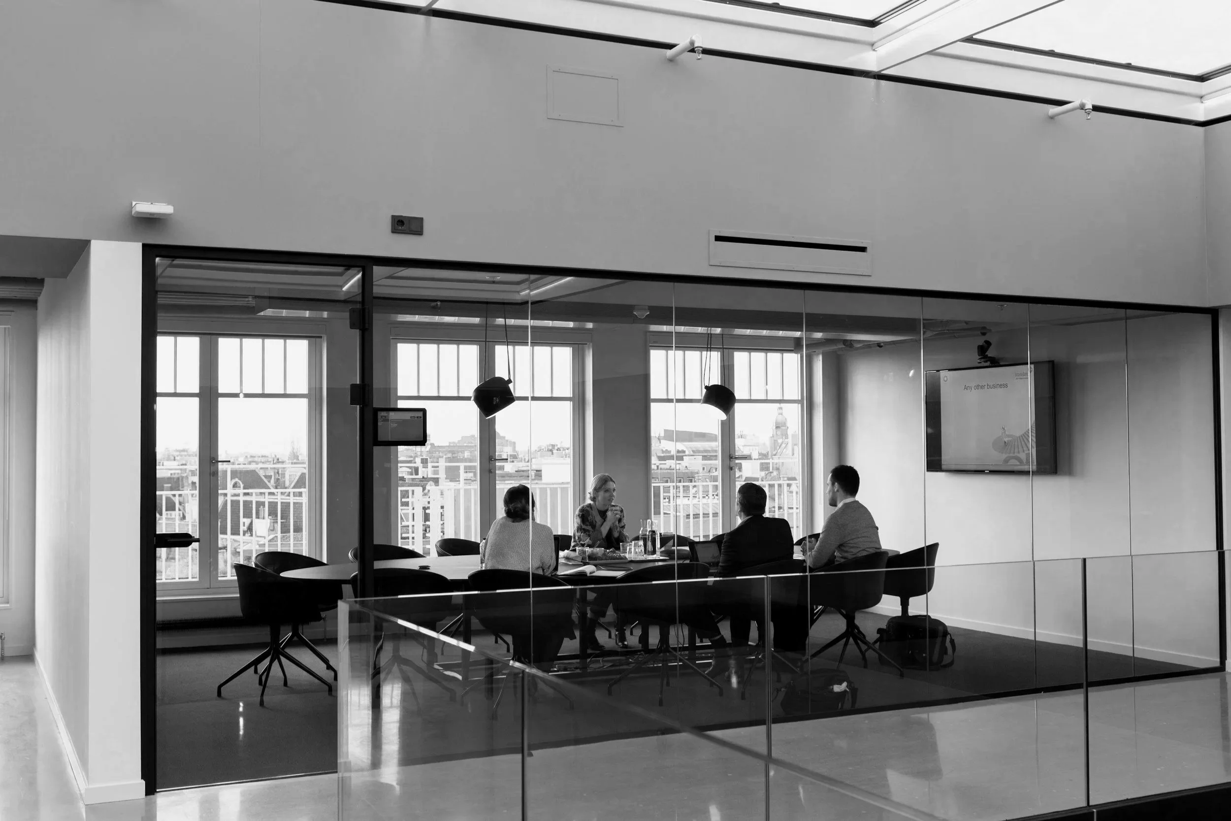 A black and white photo of a modern conference room with glass walls, five people seated around a table, and a TV screen on the wall.
