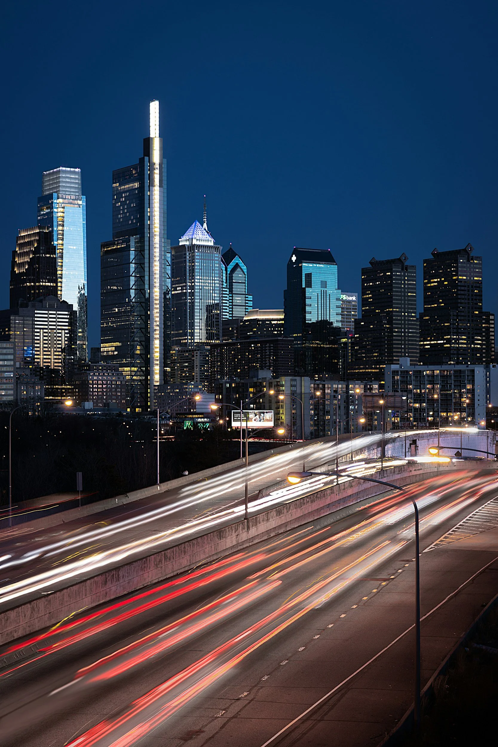 Night view of a city skyline with lit skyscrapers and a highway with light trails from moving vehicles.