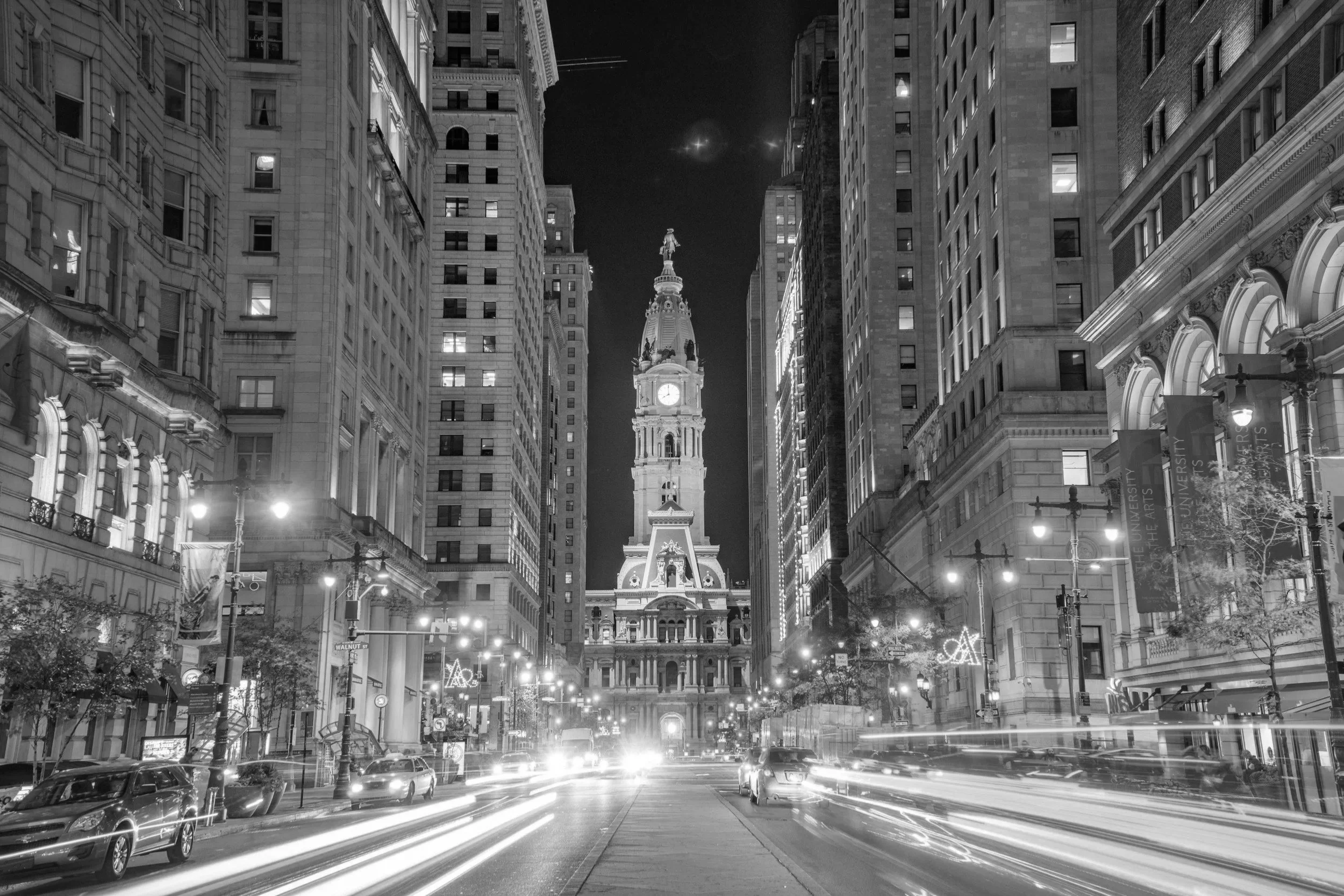 Nighttime city street with tall illuminated buildings on both sides and a historic clock tower in the distance, with light trails from moving vehicles.