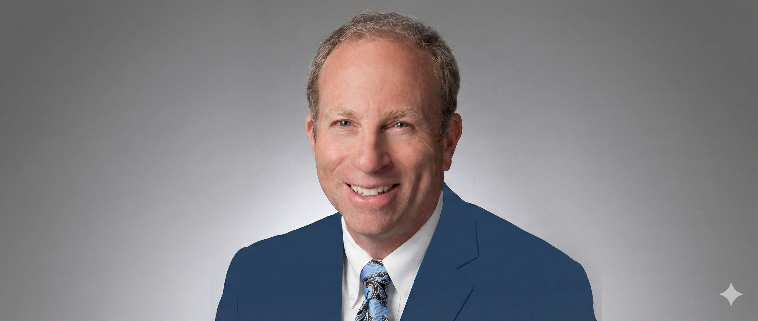 Portrait of a smiling middle-aged man with light hair, wearing a blue suit, a white shirt, and a patterned tie, set against a plain gray background.