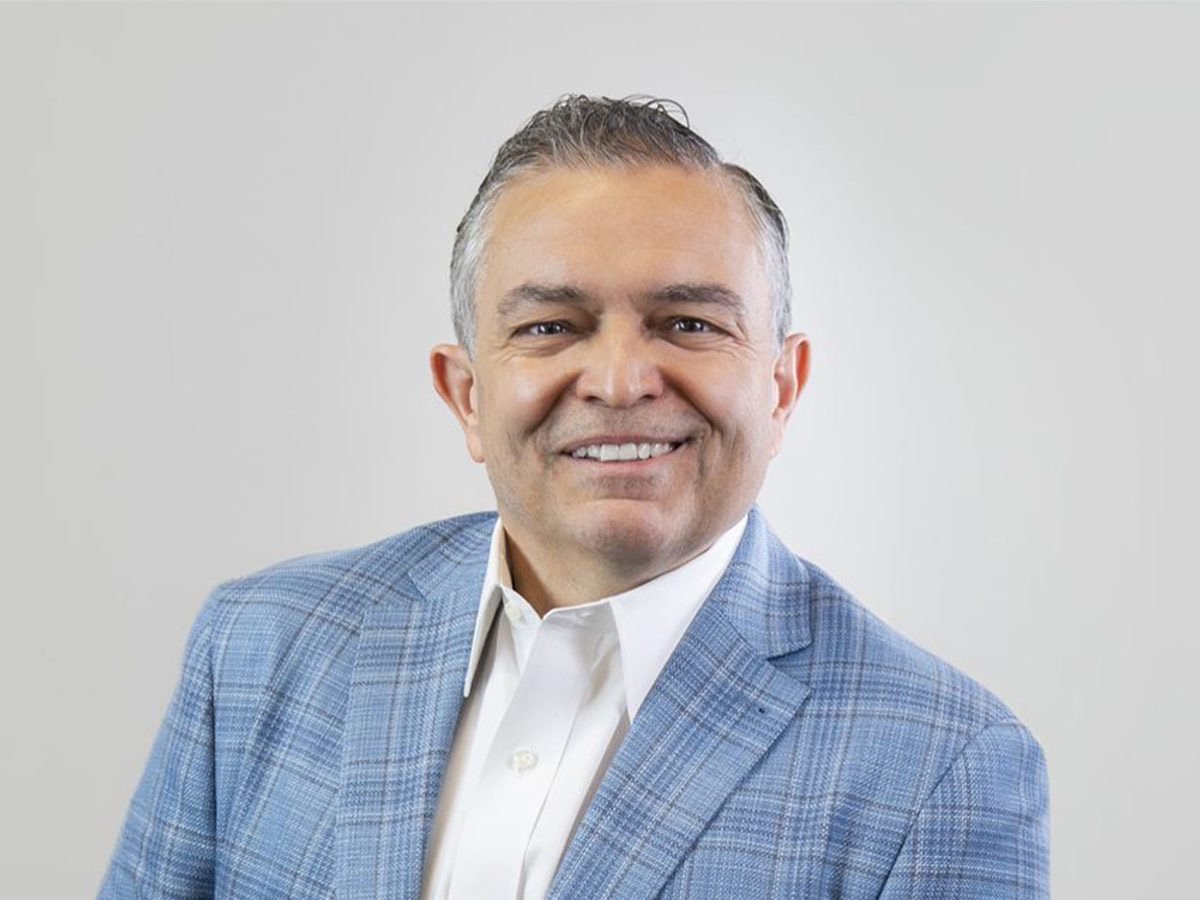 A man in a light blue checkered blazer and white dress shirt smiling at the camera against a plain gray background.
