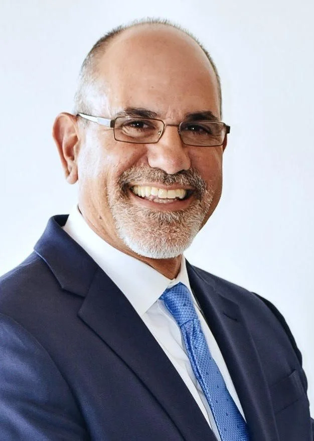 A middle-aged man with glasses, short hair, and a beard, dressed in a dark suit, white shirt, and blue tie, smiling and looking at the camera.