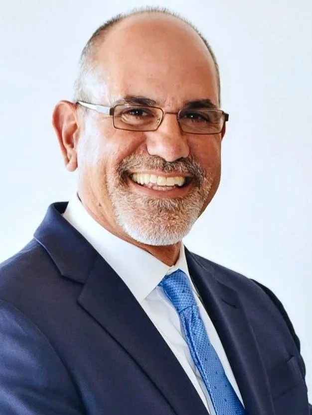 A middle-aged man with glasses and a trimmed beard, wearing a navy blue suit, a white shirt, and a blue tie, smiling at the camera.