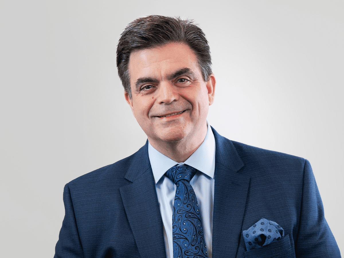 A man in a navy blue suit, light blue dress shirt, and patterned blue tie, smiling against a plain white background.