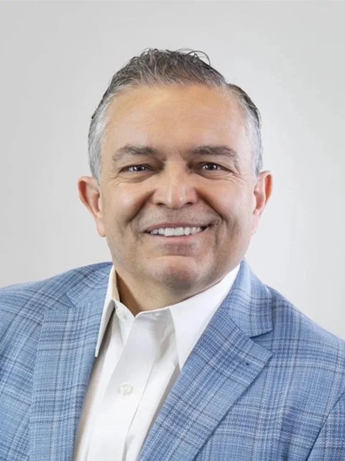 A man with short gray hair smiling, wearing a light blue plaid blazer and white shirt against a plain off-white background.