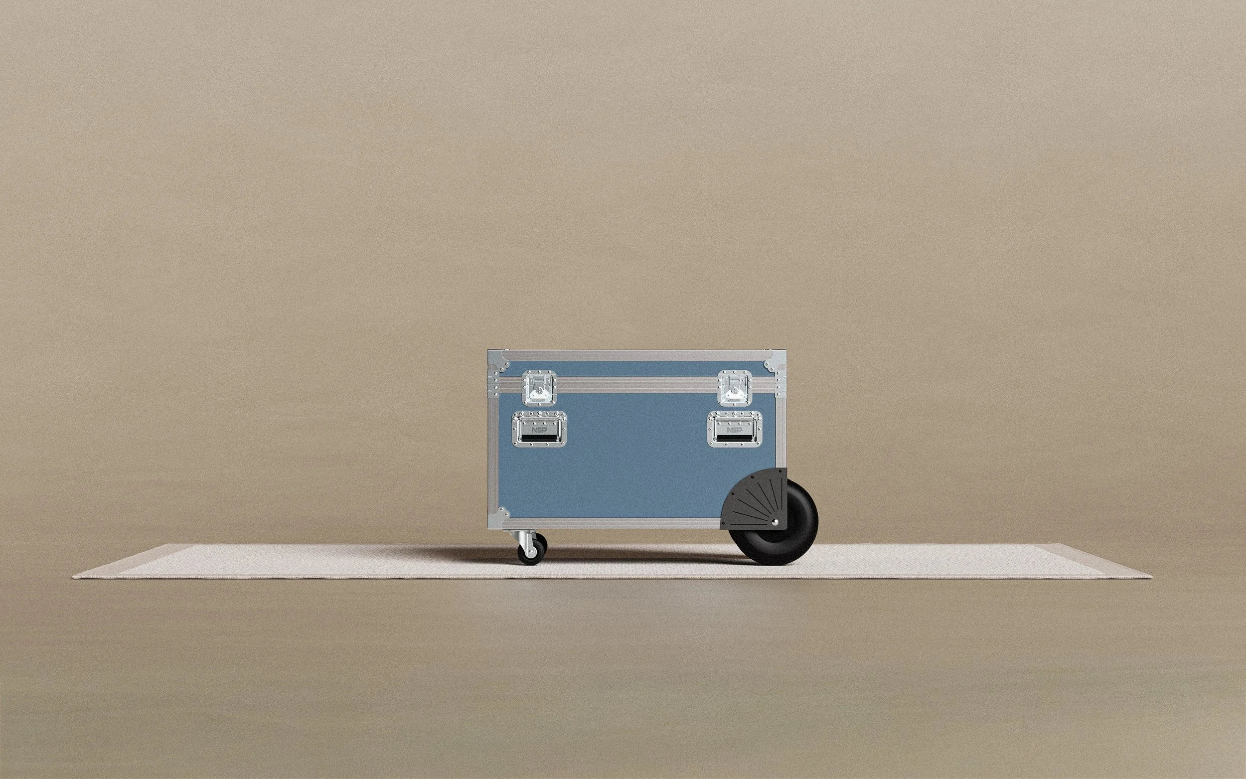 A blue equipment case with metal reinforcements and label holders, positioned on a small white rug against a beige background.
