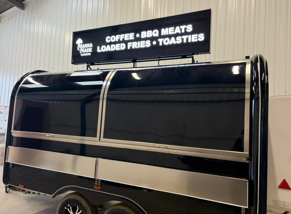 Black mobile food trailer with a digital sign on top listing offerings including coffee, BBQ meats, loaded fries, and toasties, inside a warehouse with white walls.