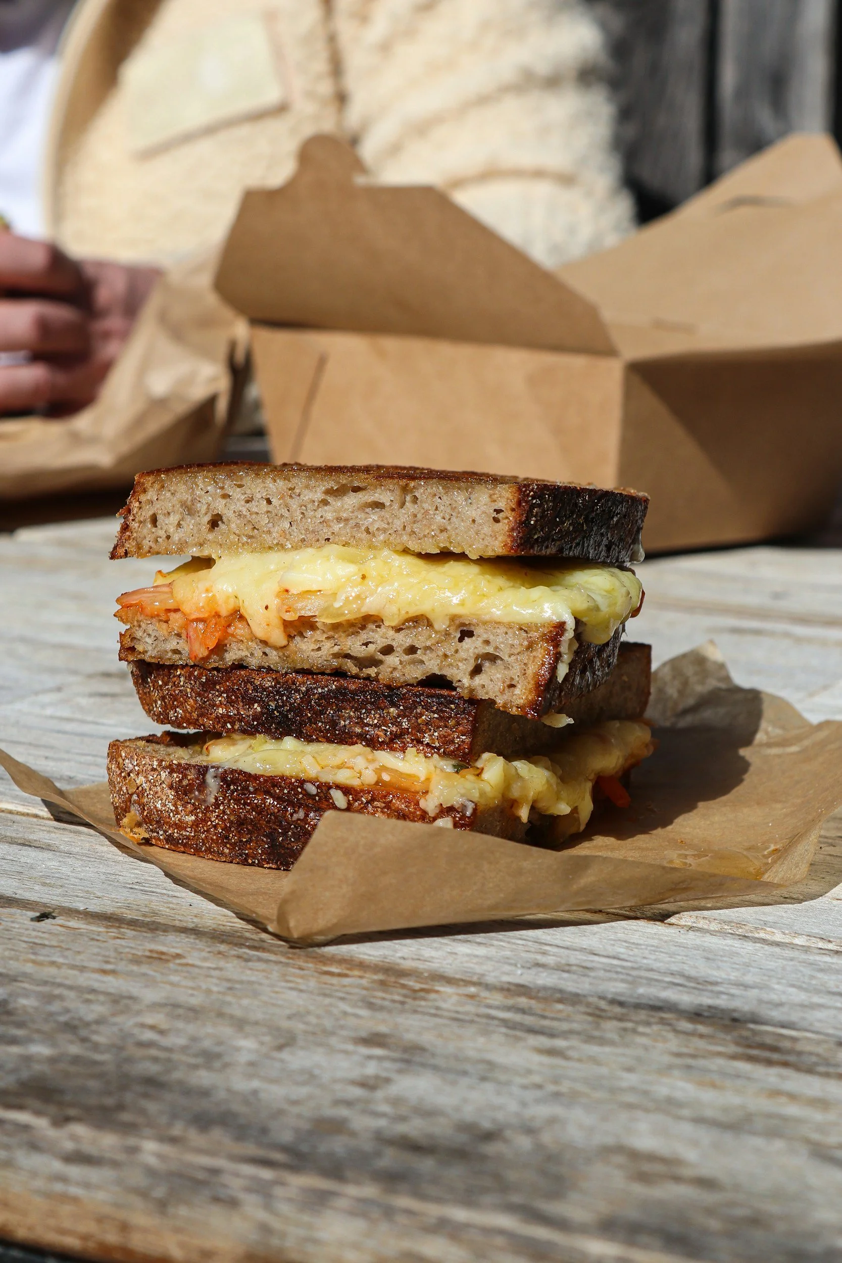 Close-up of a toasted sandwich with melted cheese and some vegetables on brown parchment paper placed on a wooden surface.