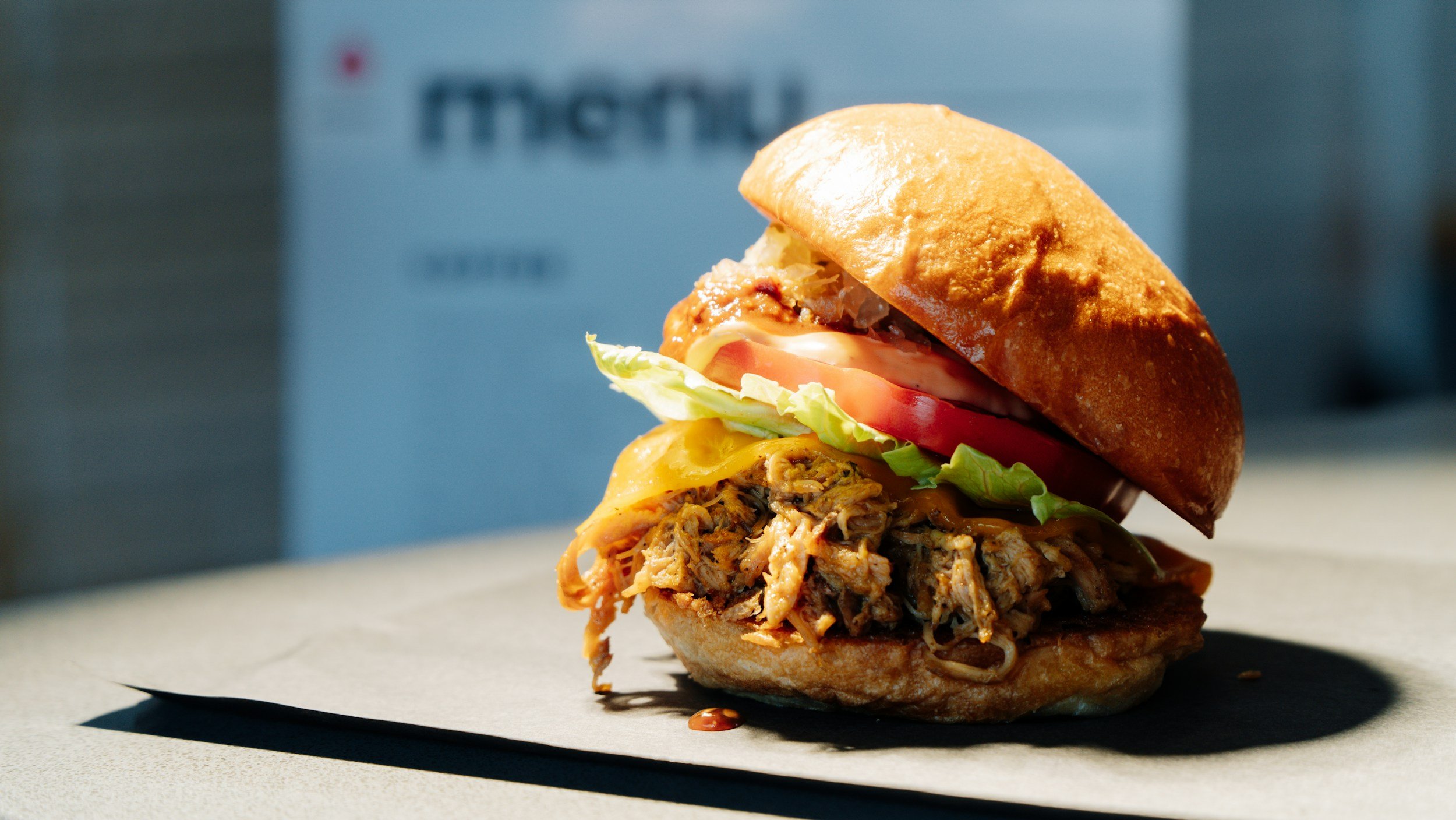 Close-up of a barbecue pulled pork sandwich with lettuce, tomato, cheese, and bun on a gray surface with a blurred background.