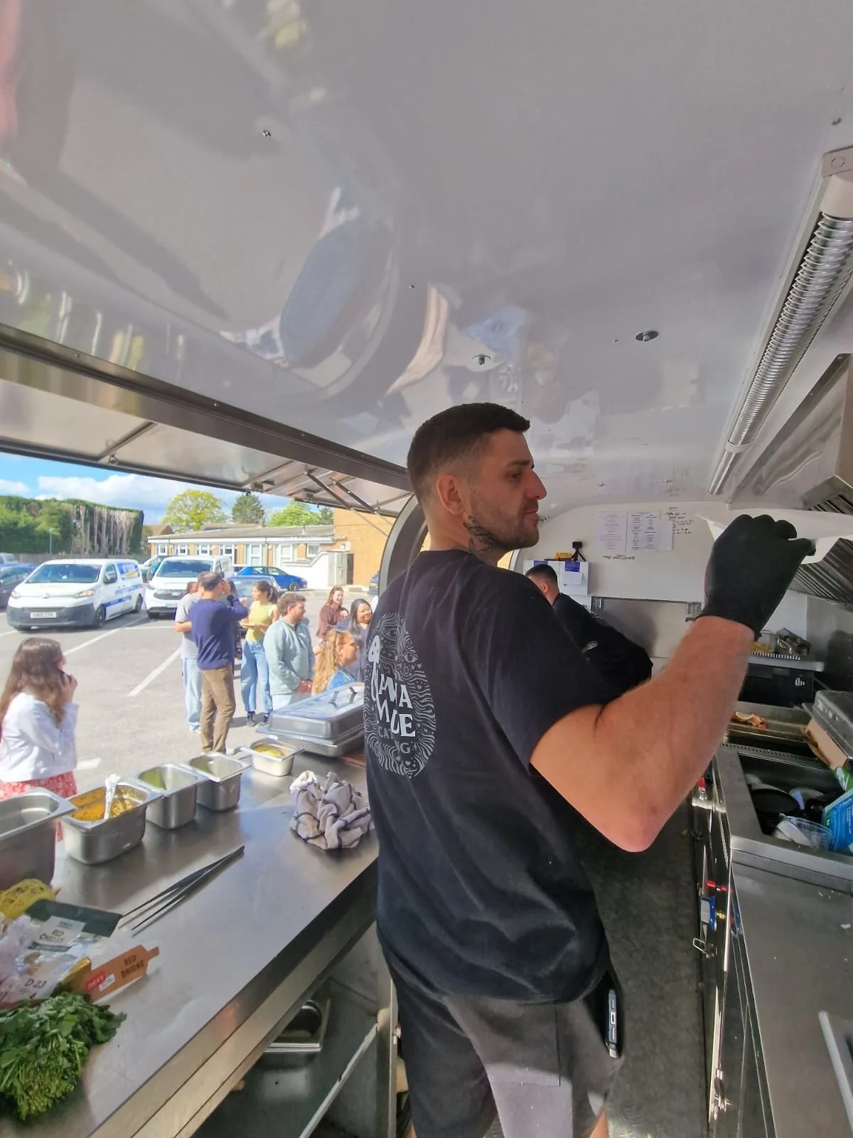 A man in a black T-shirt and black gloves is working inside a food truck. Outside the truck, a line of people is waiting, with some talking and others waiting. The food truck has a stainless steel counter with ingredients and utensils, and behind the man, there are menu papers on the wall.