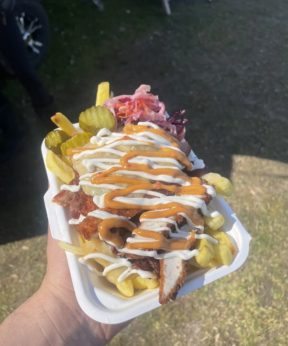 A hand holding a fried chicken sandwich topped with pickles, onions, coleslaw, and drizzled with mayonnaise and hot sauce, served in a white foam tray.