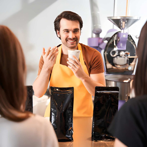 Man in a yellow apron holding a cup, smiling during a coffee tasting session with two women at a coffee roastery.