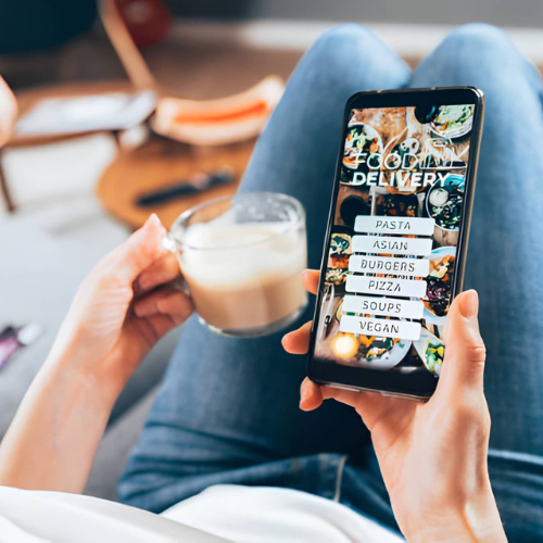 Person holding a smartphone displaying a food delivery app with options for pasta, Asian, dinners, pizza, soups, and vegan, while holding a glass of latte in the other hand, sitting with legs crossed.