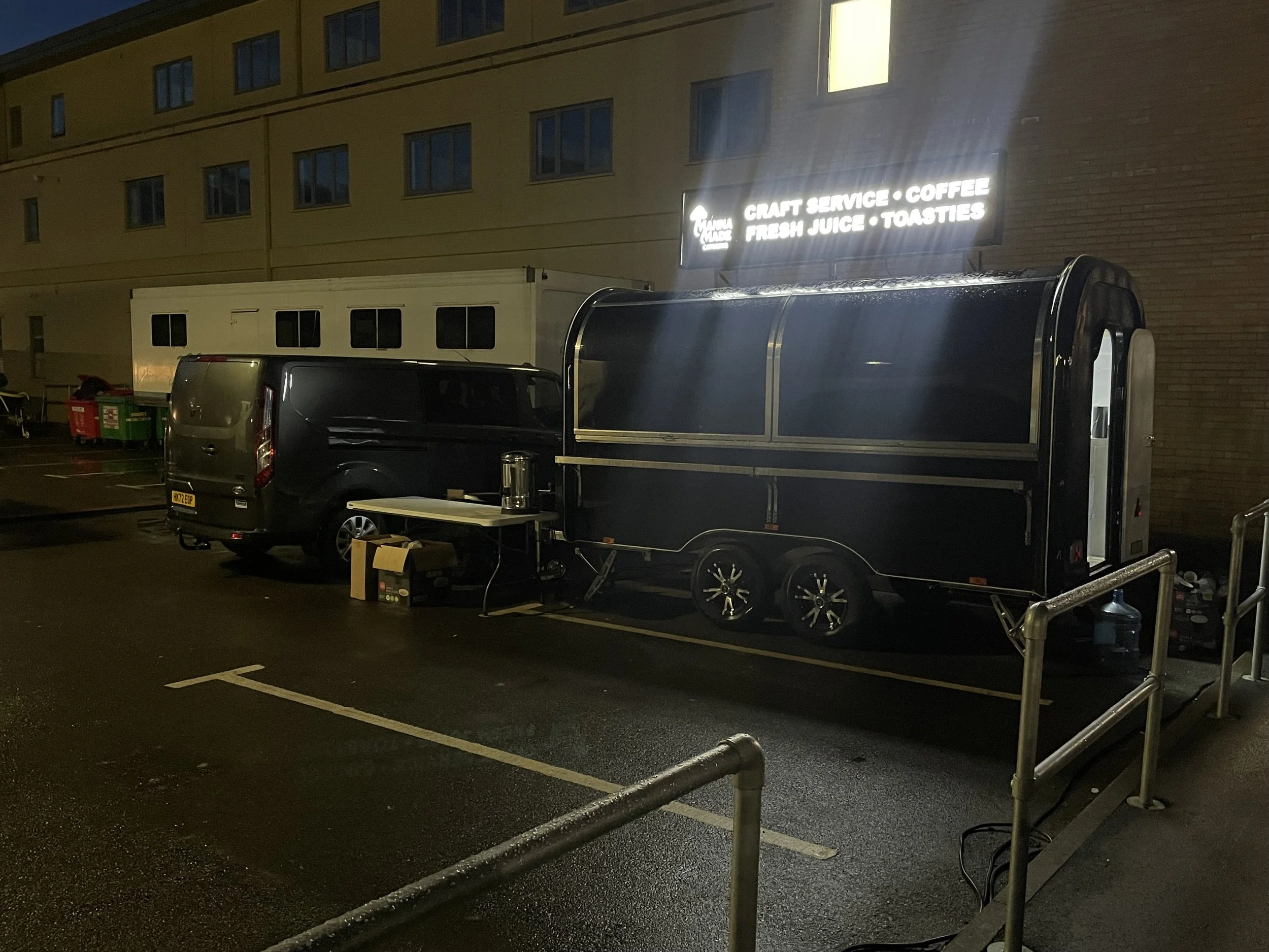 A food truck parked in a lot at night, with a black van beside it. The food truck has an illuminated sign that reads 'Craft Service, Coffee, Fresh Juice, Toasties'. There are some boxes and a coffee pot on a small table near the truck.