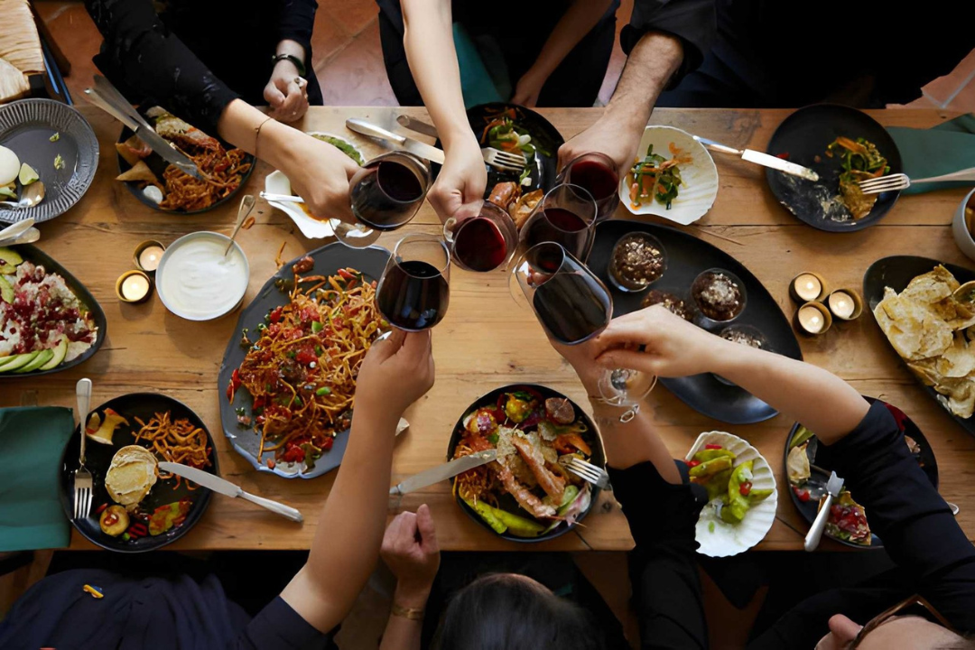 People enjoying a meal and raising glasses of red wine in a toast at a dinner table with various dishes and desserts.