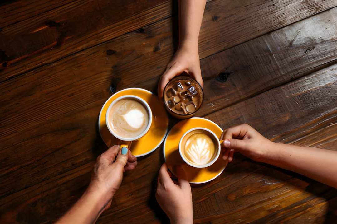 Three people holding coffee cups, two with latte art, and a glass of iced coffee with ice cubes, on a wooden table.