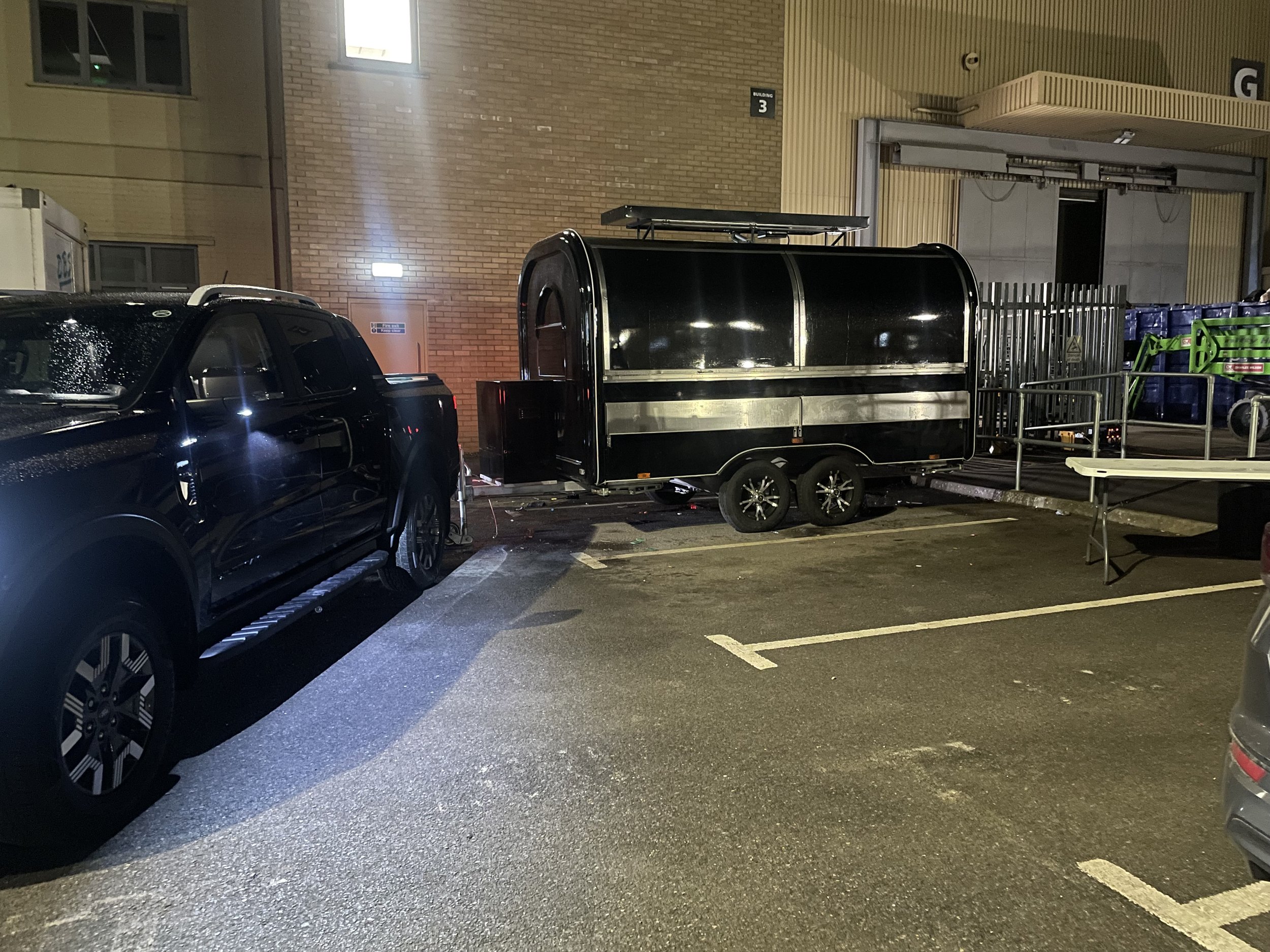 A black pickup truck parked next to a small black trailer in a parking lot at night, with a brick building and some green equipment in the background.