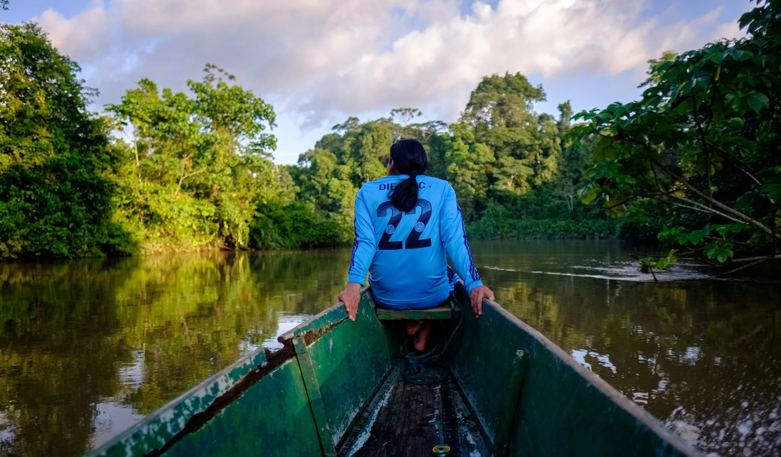 A person wearing a blue sports jersey with the number 22 sitting at the front of a boat on a river, surrounded by lush green trees under a cloudy sky. Amazon jungle boat ride.