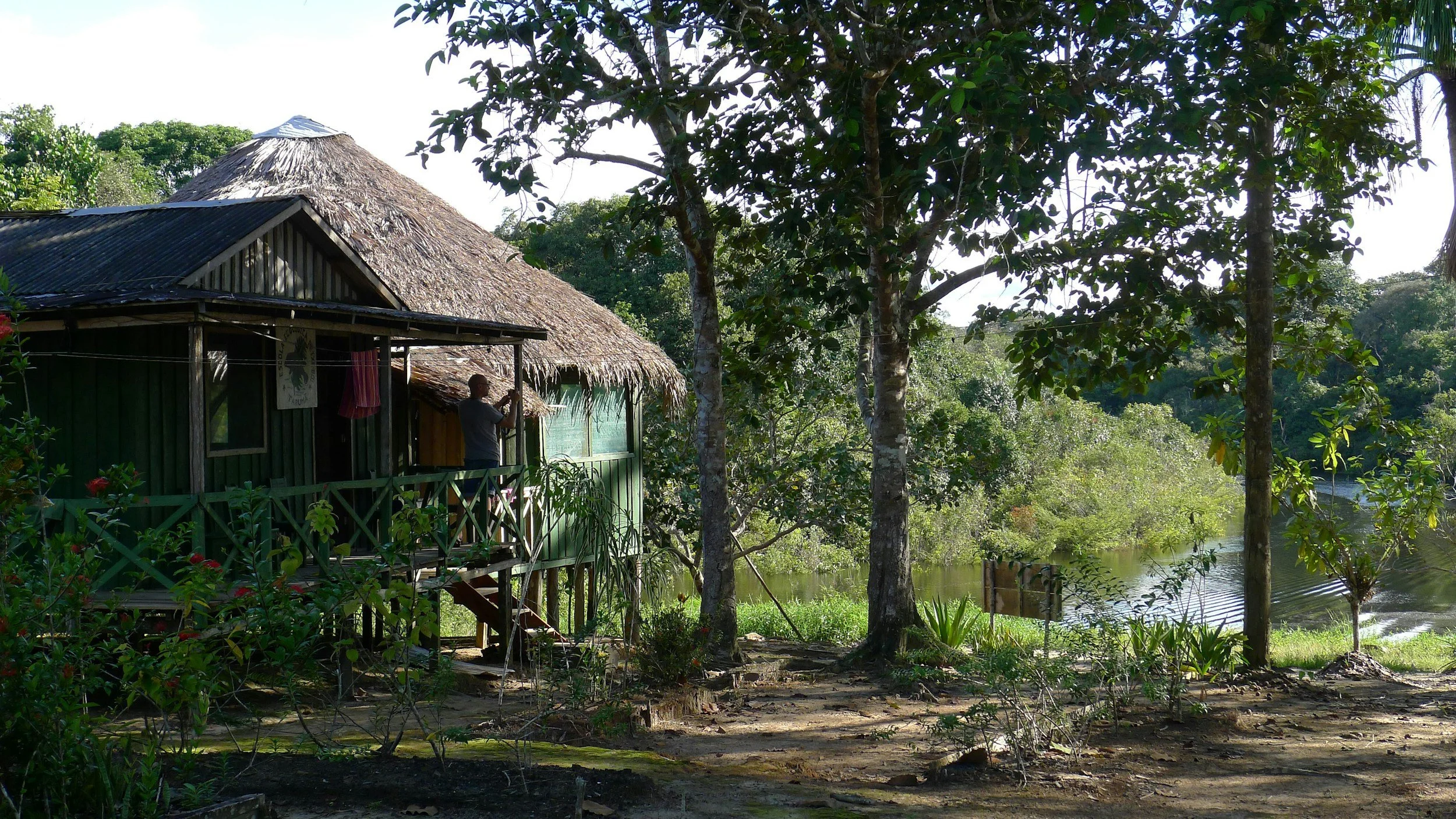 A person taking a photo on a porch of a rustic house made of wood and bamboo, surrounded by lush trees and overlooking a river.