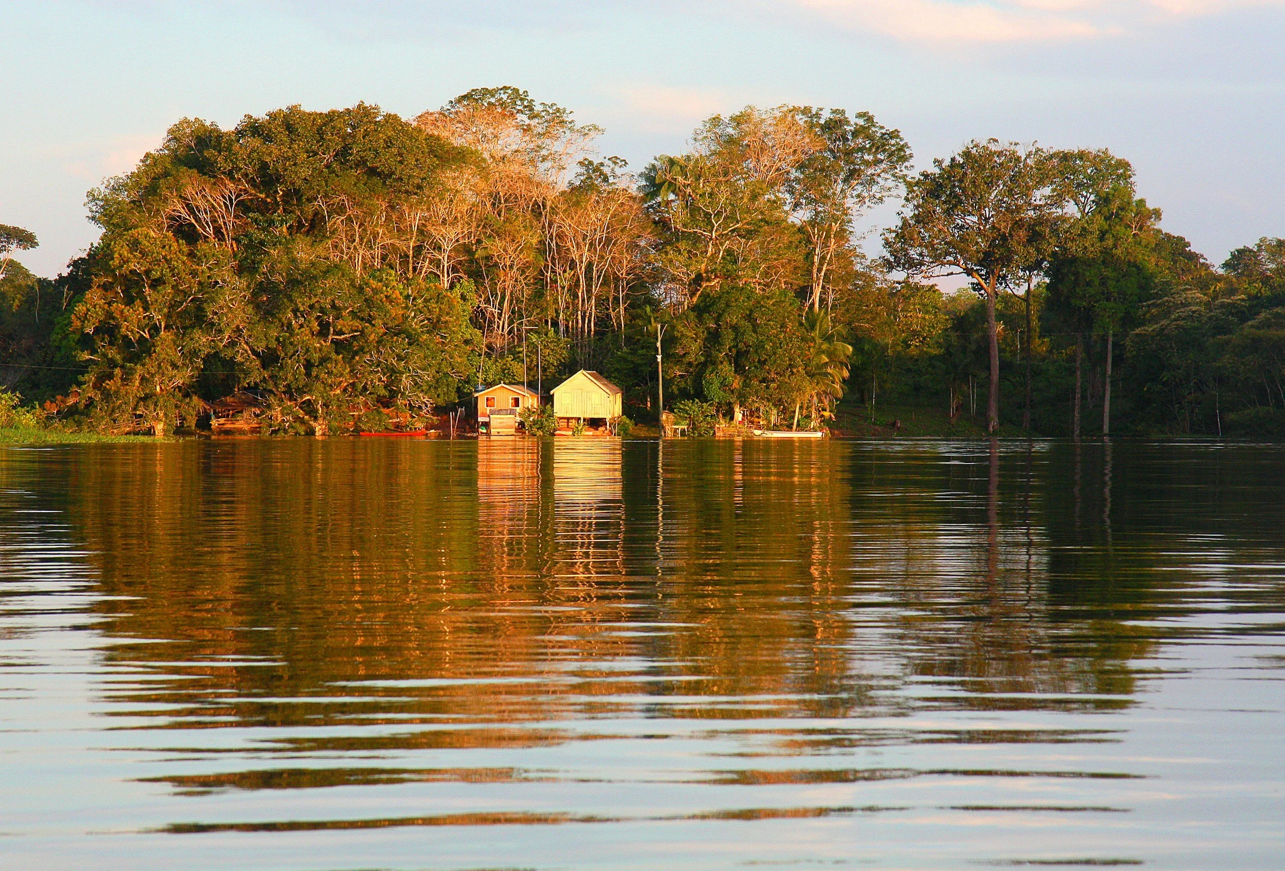 A tranquil view of a river reflecting a small, green house and trees on the riverbank at sunset.