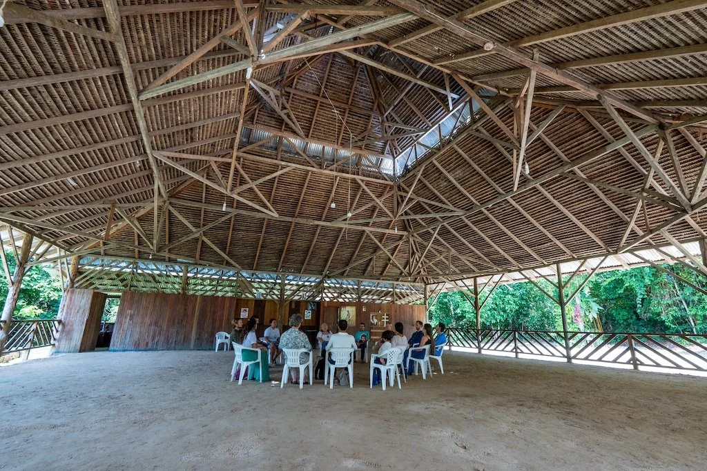 People sitting in a circle on plastic chairs under a large open wooden pavilion. Indigenous medicine ceremony, healing, community, spirituality.