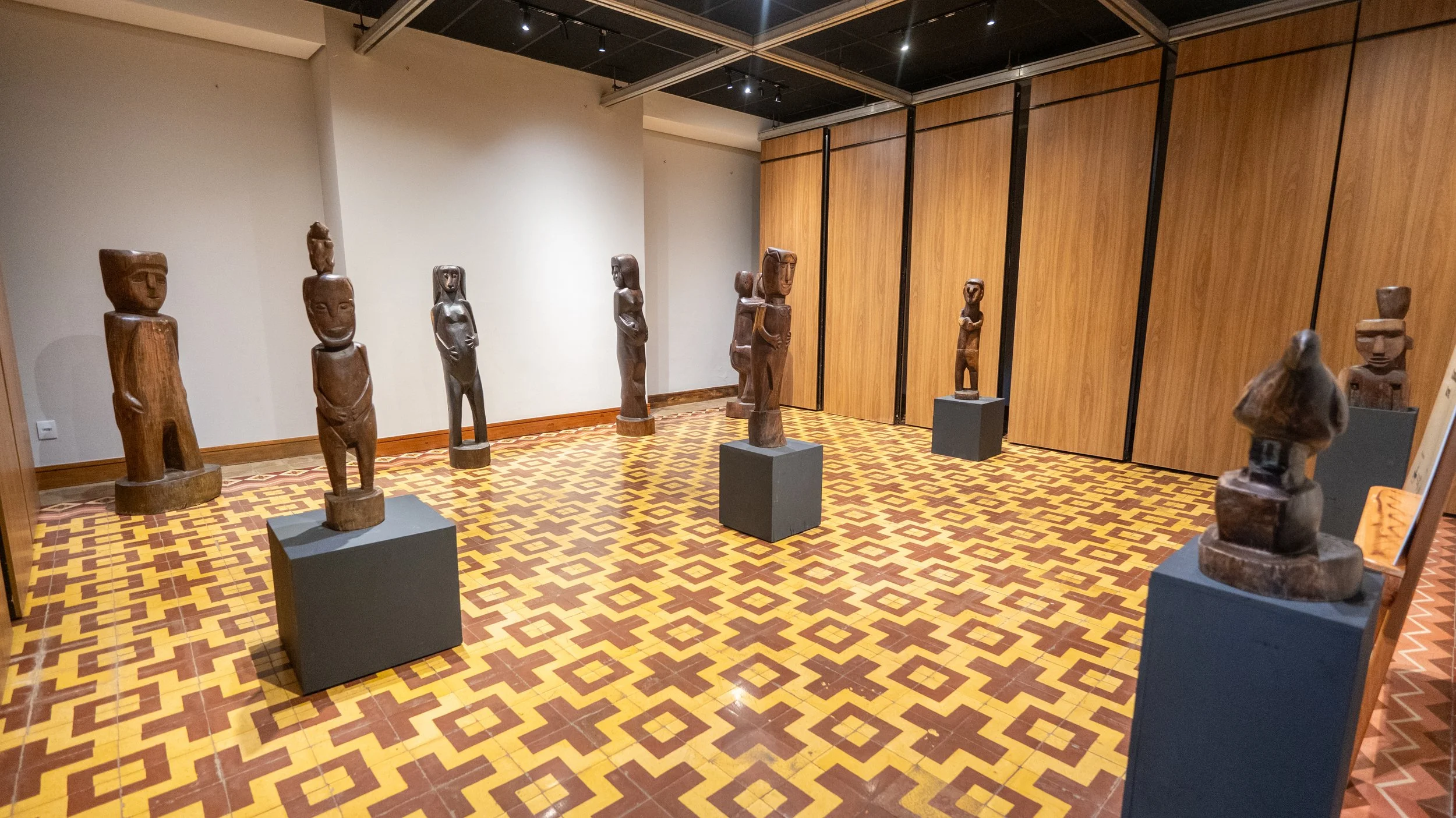 Museum exhibit with nine wooden sculptures of human figures on black pedestals, arranged in a gallery with patterned tile floor and wooden-paneled walls.
