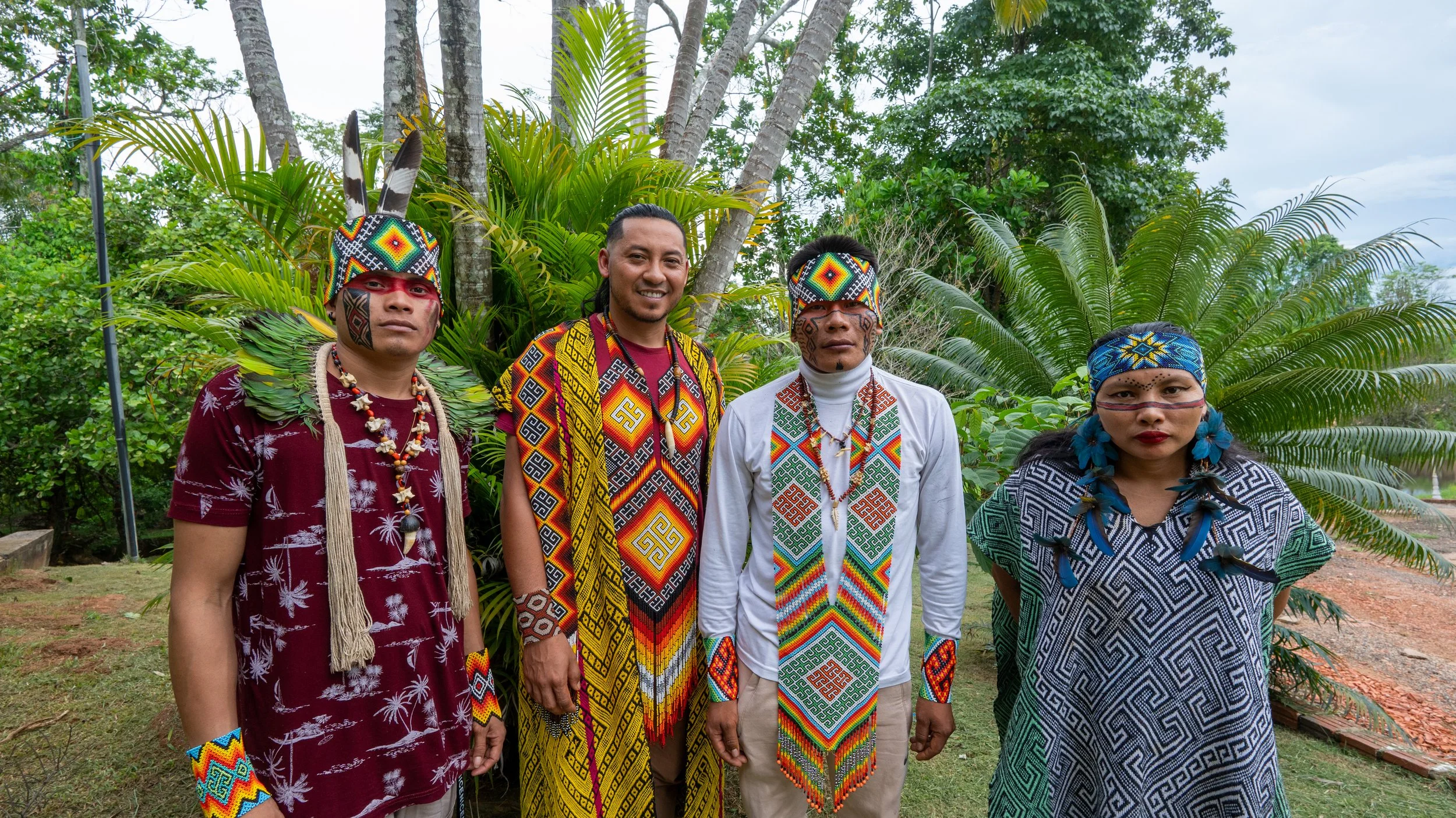 Four people standing outdoors, dressed in traditional indigenous clothing with vibrant beadwork and patterns, surrounded by lush green plants and trees. Huni Kuin tribe with traditional face paint.