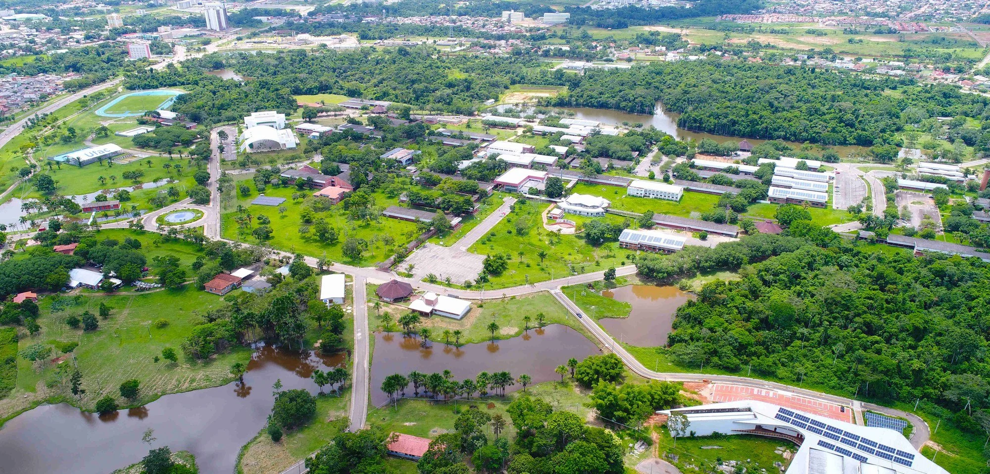 Aerial view of a university campus with green lawns, buildings, water bodies, trees, parking lots, and sports fields in a lush, green landscape.
