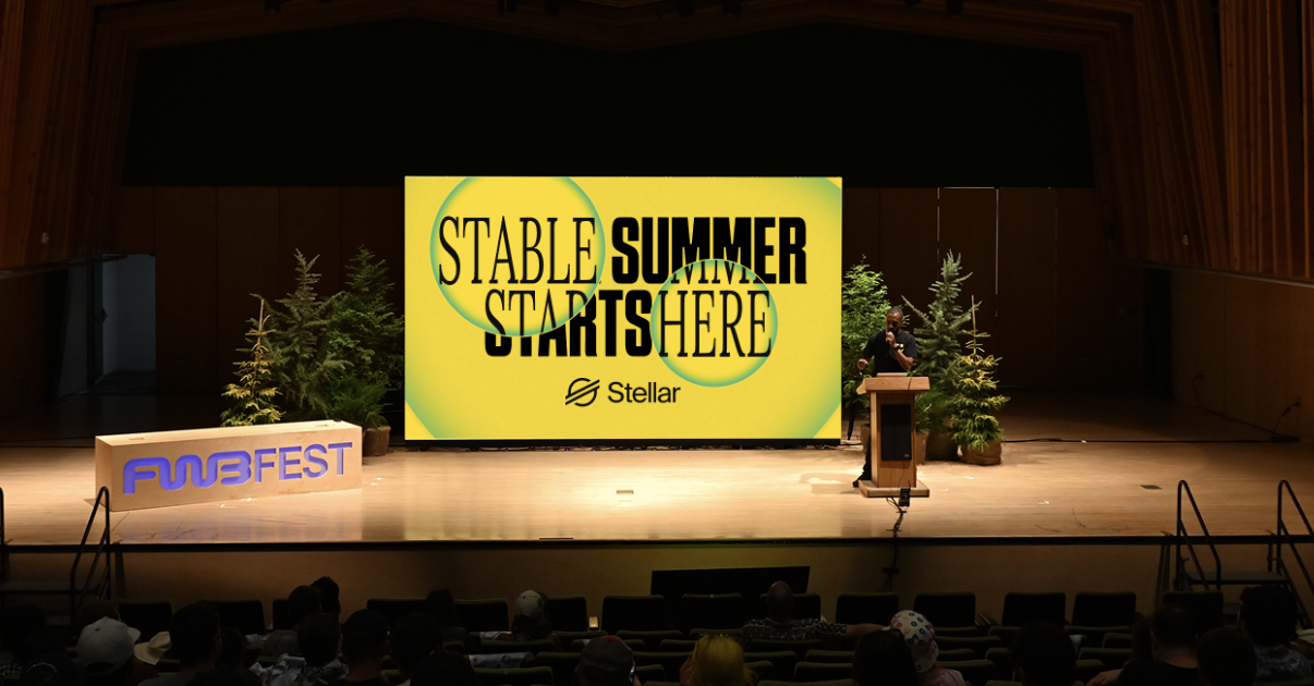 A stage set for a conference with a large yellow screen displaying the text 'STABLE SUMMER STARTS HERE' and the 'Stellar' logo. There are small trees on both sides of the screen, a wooden podium with a person speaking, and a sign on the left that reads 'FUB FEST.' Audience members are seated in front.