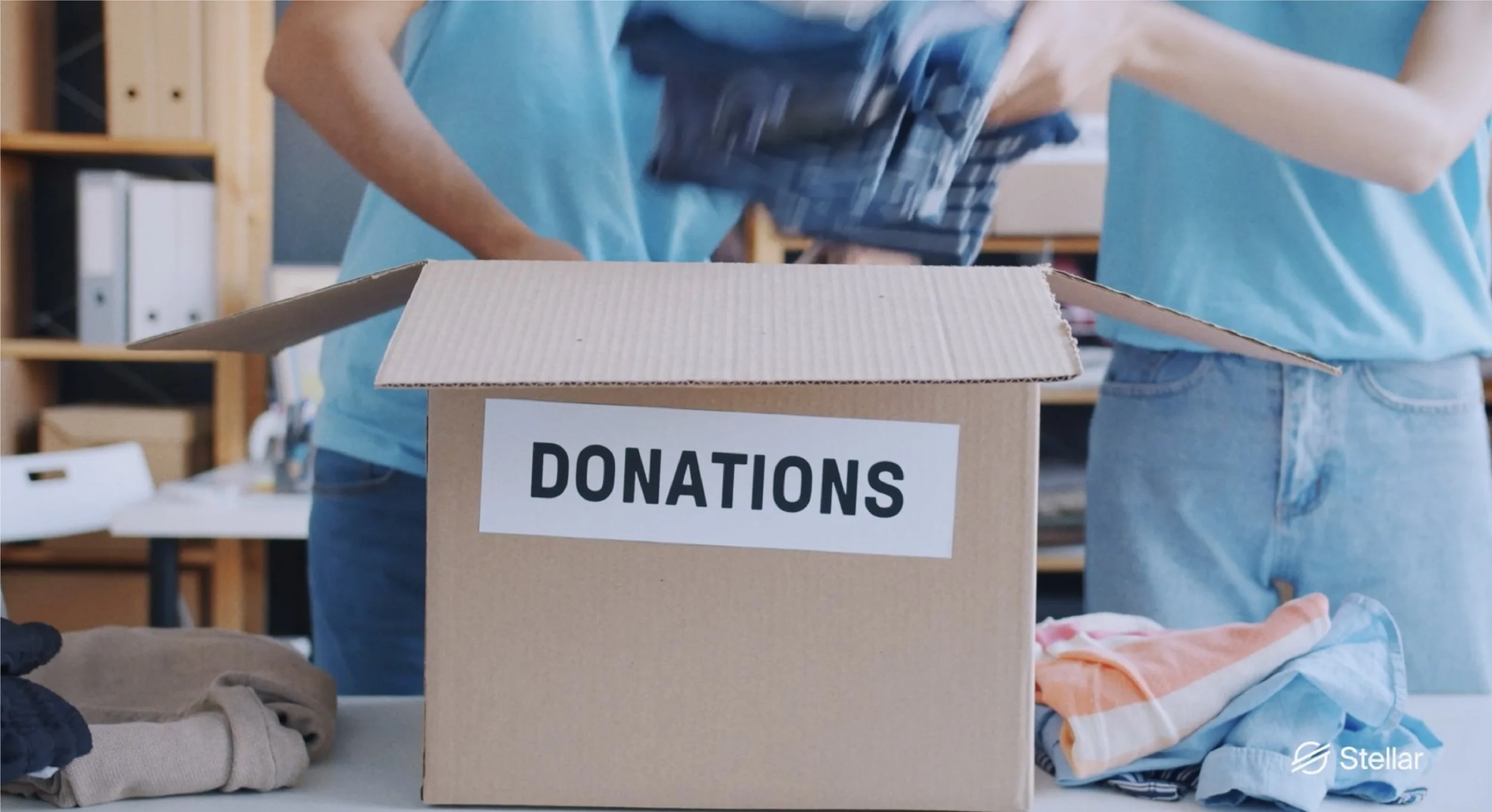People packing donations into a cardboard box labeled "DONATIONS" on a table, with clothes nearby.