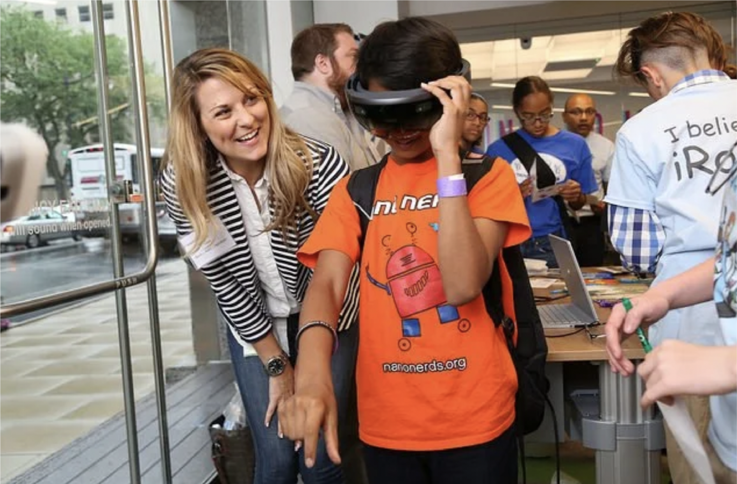 A group of people at a technology event, with a woman in a striped blazer smiling and leaning toward a person wearing an orange T-shirt and virtual reality headset, surrounded by other attendees.