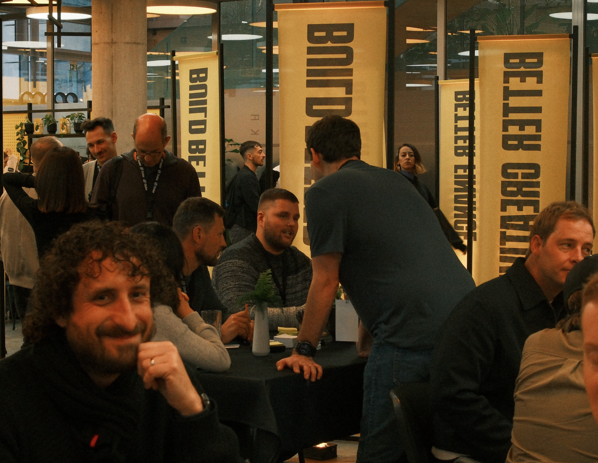 People socializing at an indoor event with yellow banners displaying ‘BETTER BOLD’ and ‘BUILDING BETTER’ behind them. Several individuals are seated at tables, engaged in conversation, with some standing and chatting in the background.